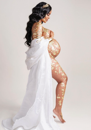 Artistic maternity photo with flowing white fabric and gold body art against light backdrop.
