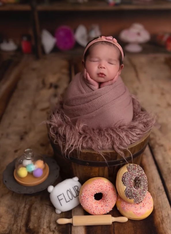 A sleeping newborn wrapped in mauve fabric posed in a wooden barrel with decorative donuts and treats.