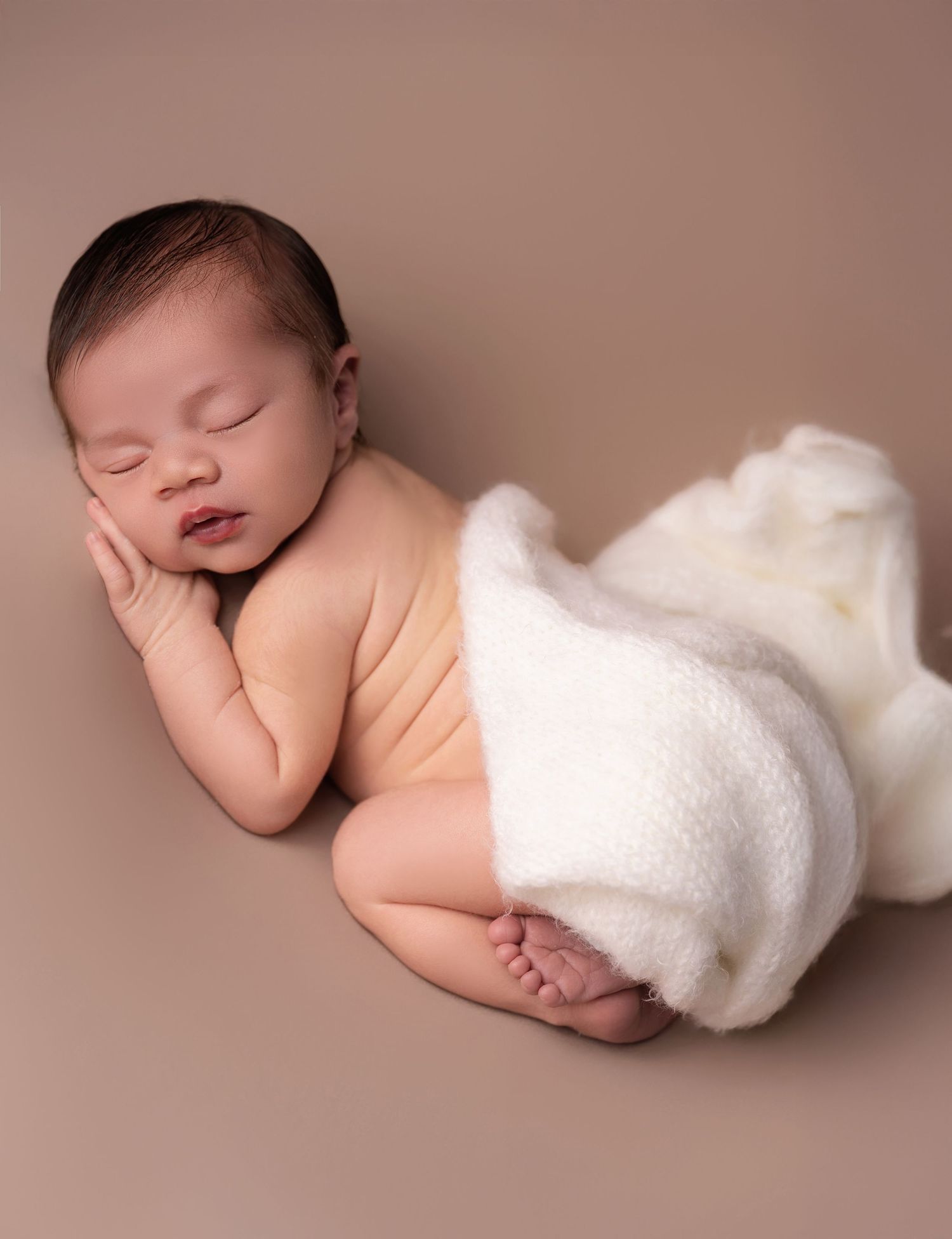 Peaceful newborn baby sleeping curled up on a beige backdrop, wrapped in a soft white knit blanket.