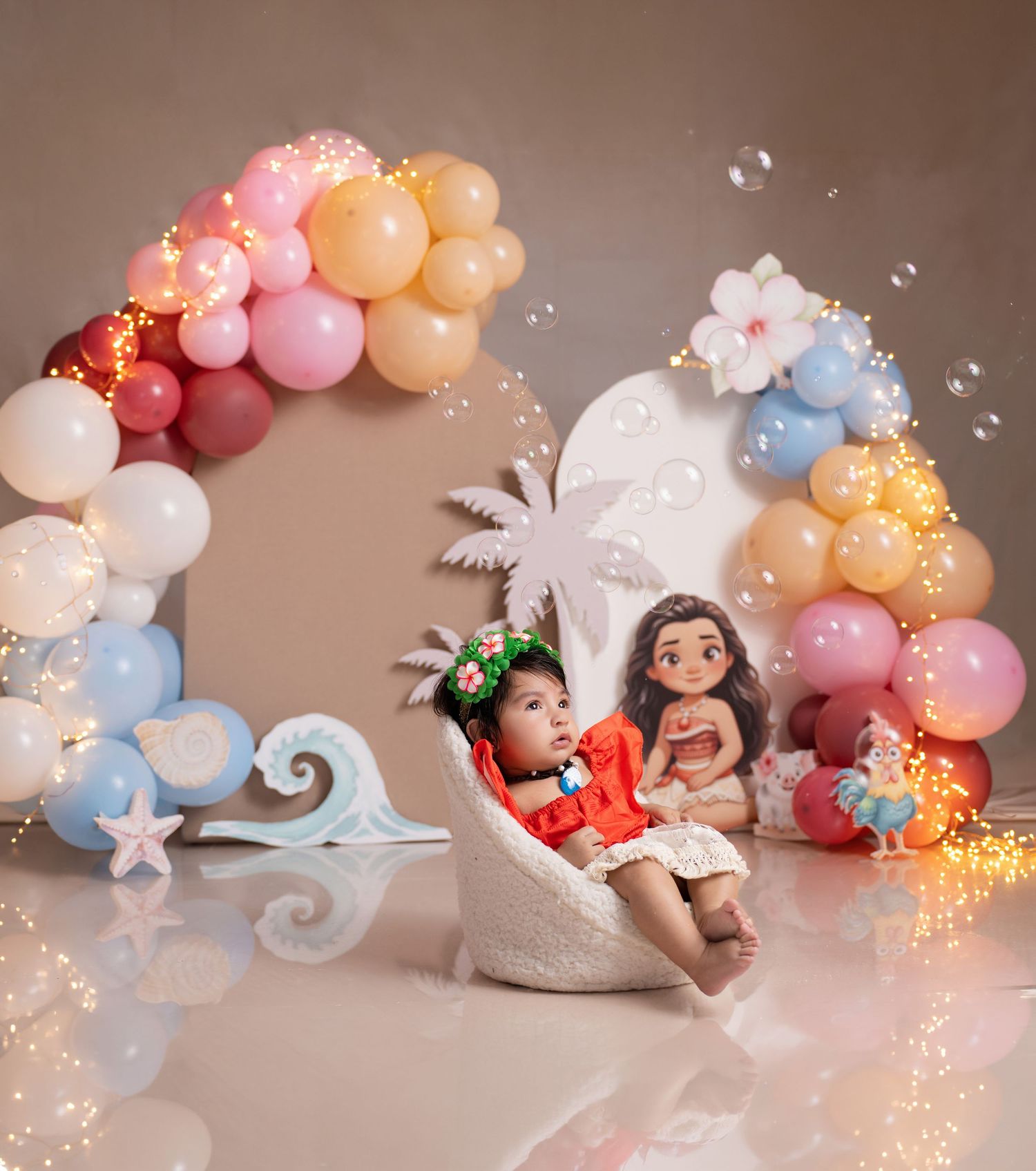 Baby sits in white chair holding Moana doll surrounded by colorful balloon arch with ocean-themed decorations and fairy lights.