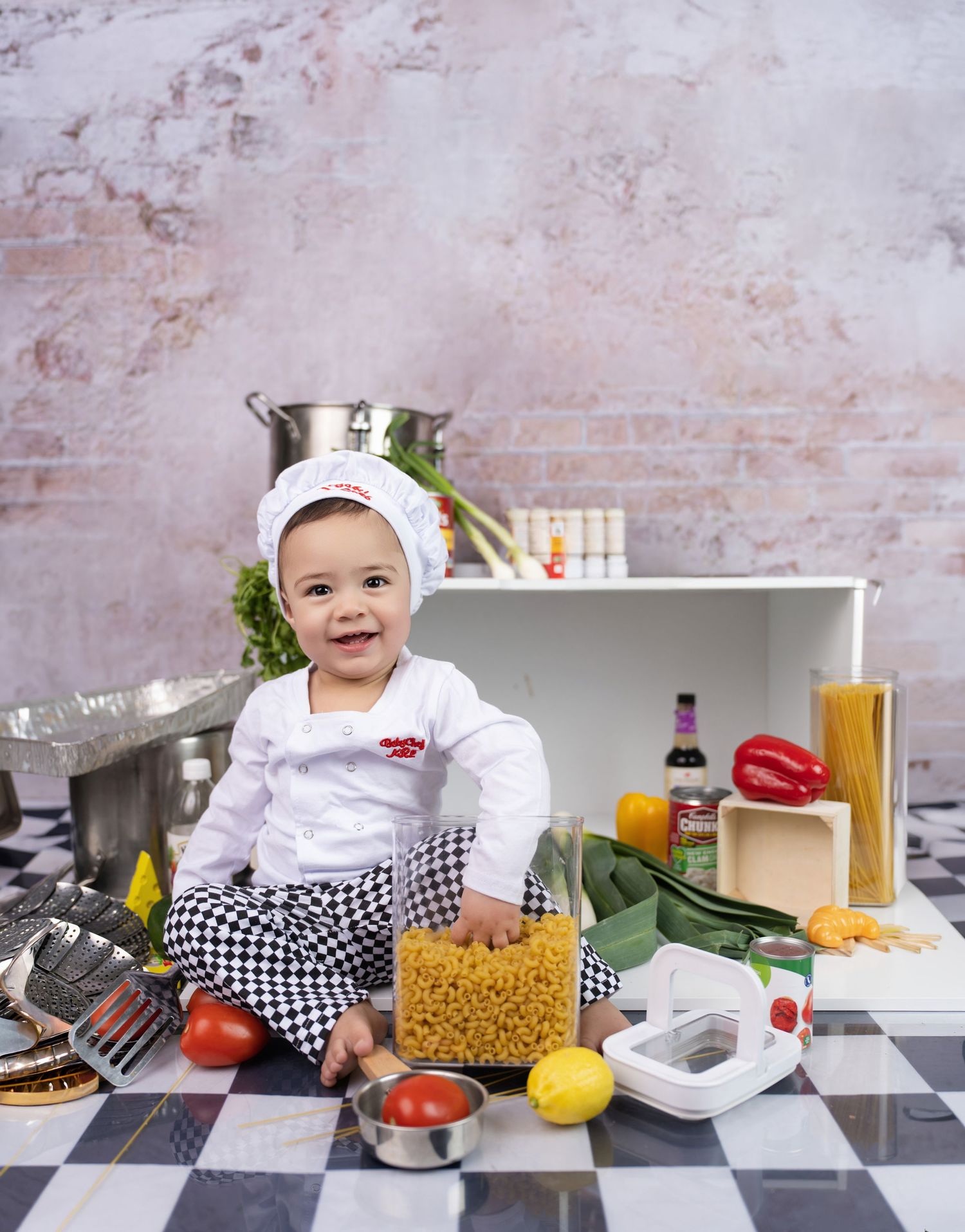 Adorable baby dressed as chef with white hat sitting among fresh vegetables and pasta ingredients on kitchen floor.