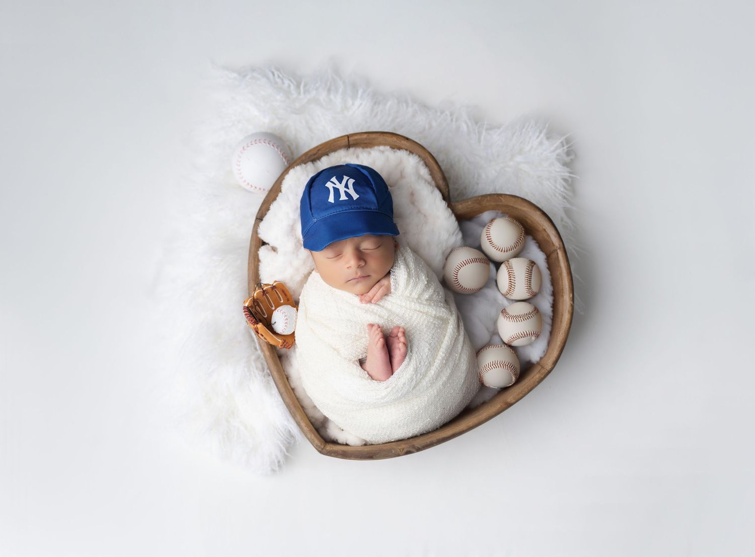 Newborn baby wrapped in white swaddle wearing NY Yankees cap sleeping in heart-shaped wooden bowl with baseballs.