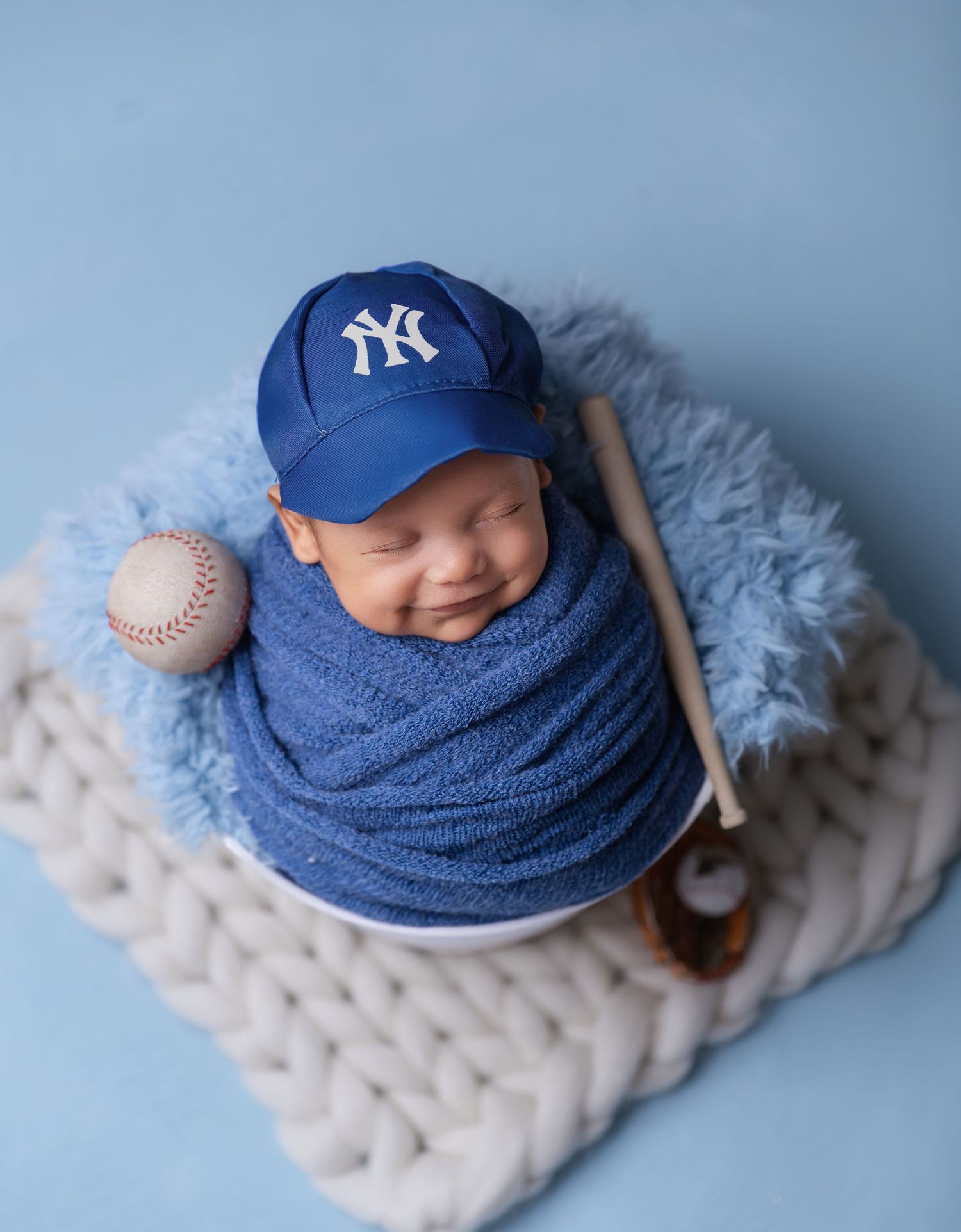 Smiling newborn baby wrapped in blue, wearing a NY Yankees cap, posed with a baseball and bat in a bowl.