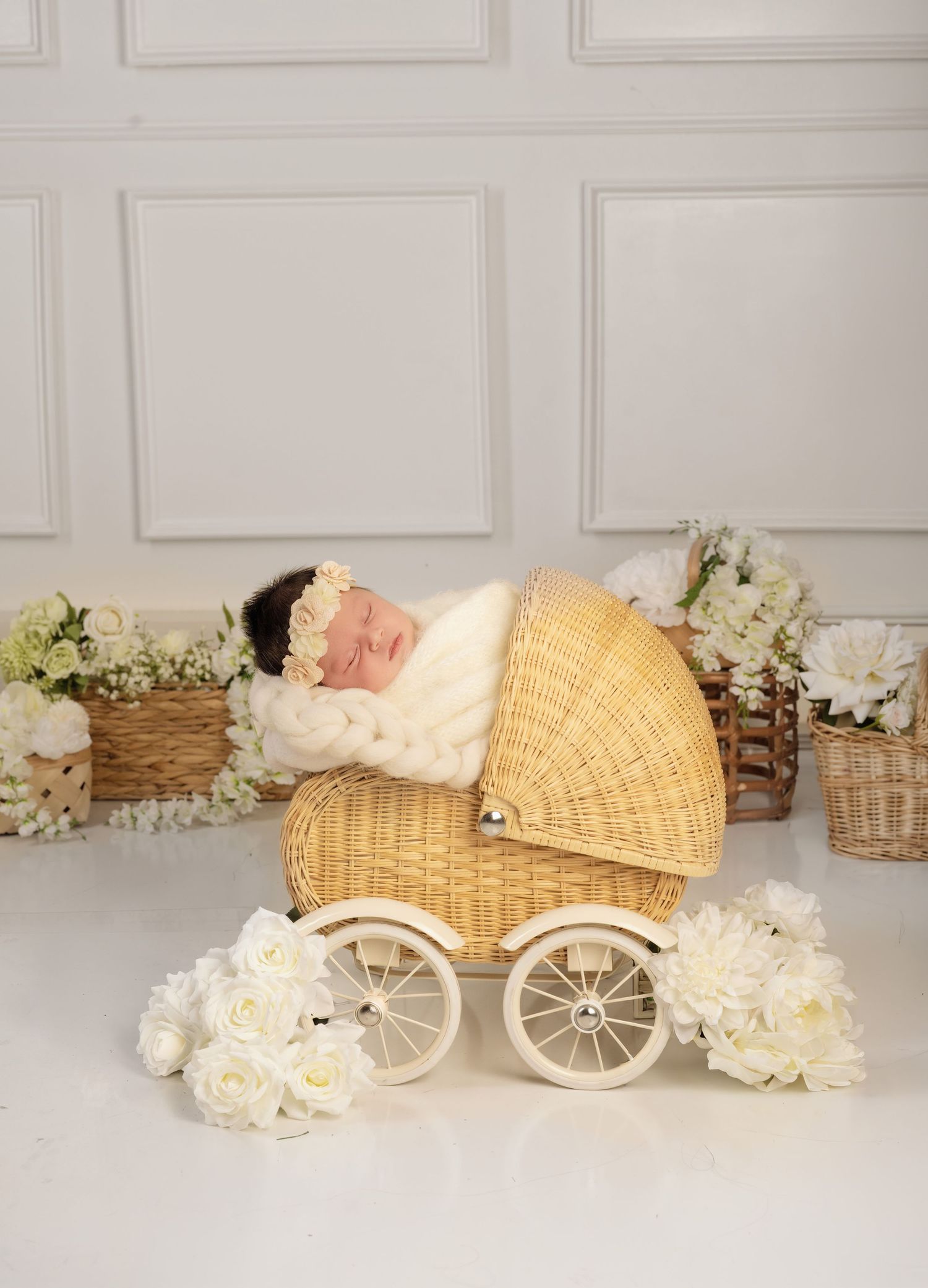 Newborn baby sleeping peacefully in a vintage wicker pram surrounded by white roses and floral arrangements.