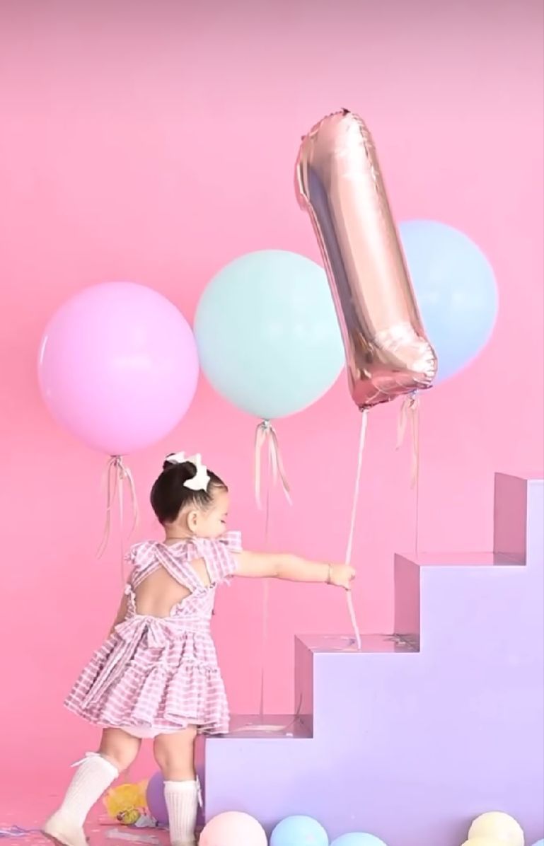Small child climbing pink stairs holding large gold number one balloon with pastel decorations.