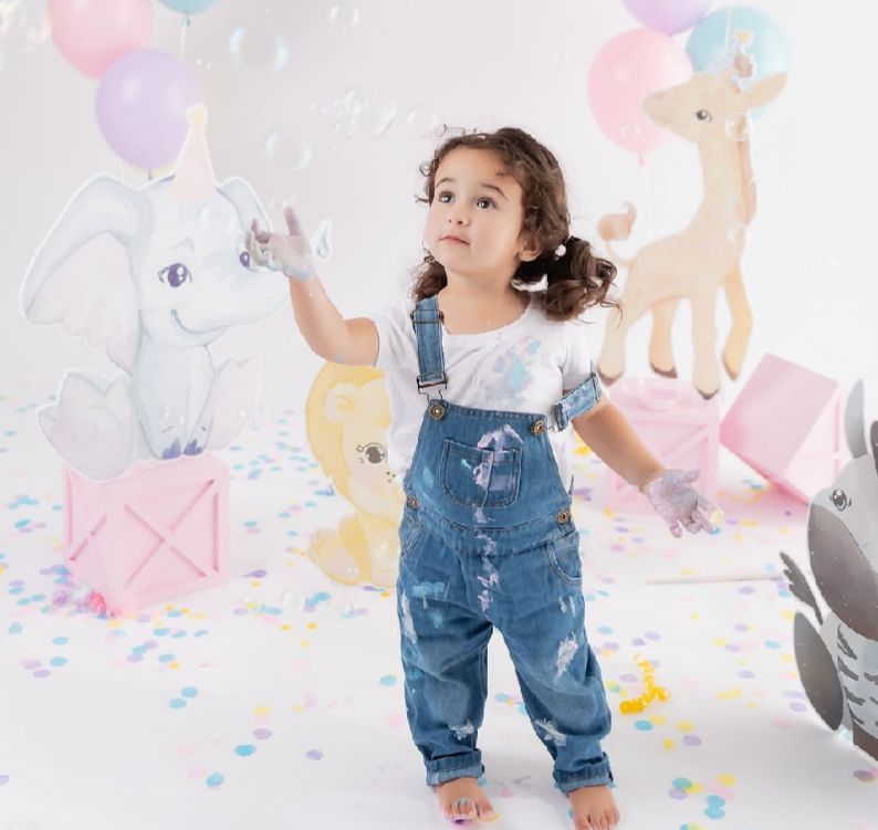 A child in denim overalls plays in a whimsical pastel-colored setting with balloons and illustrated animal decorations.