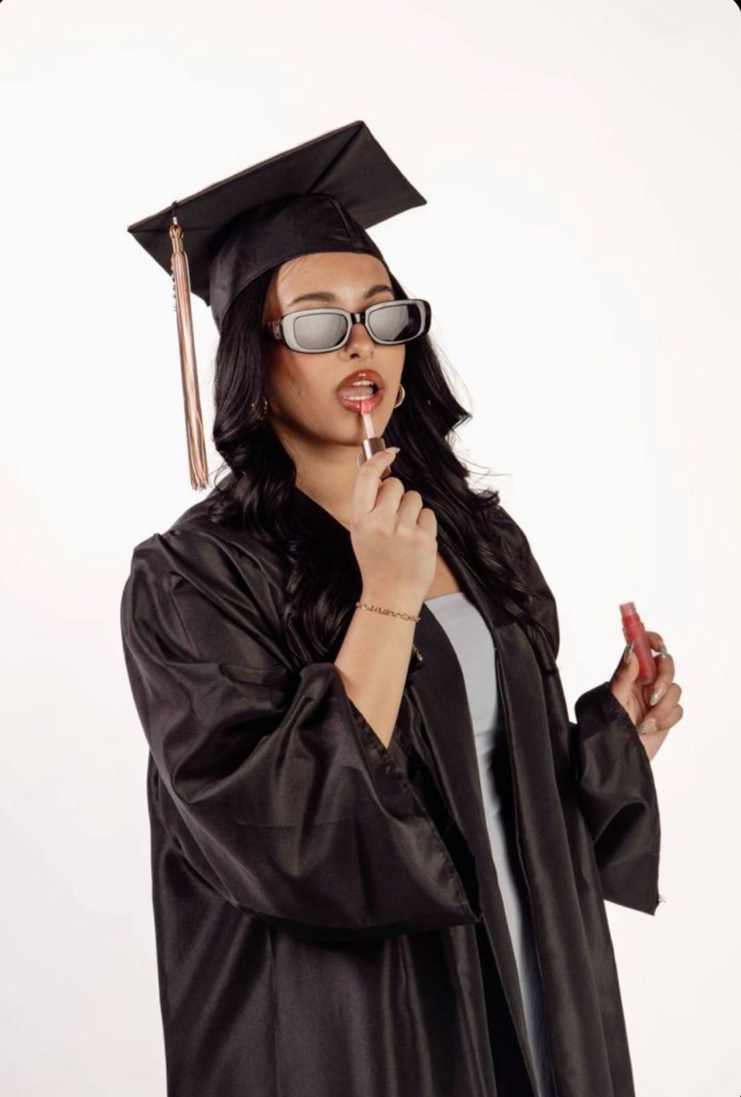 Graduate in black cap and gown wearing glasses strikes thoughtful pose against white background.