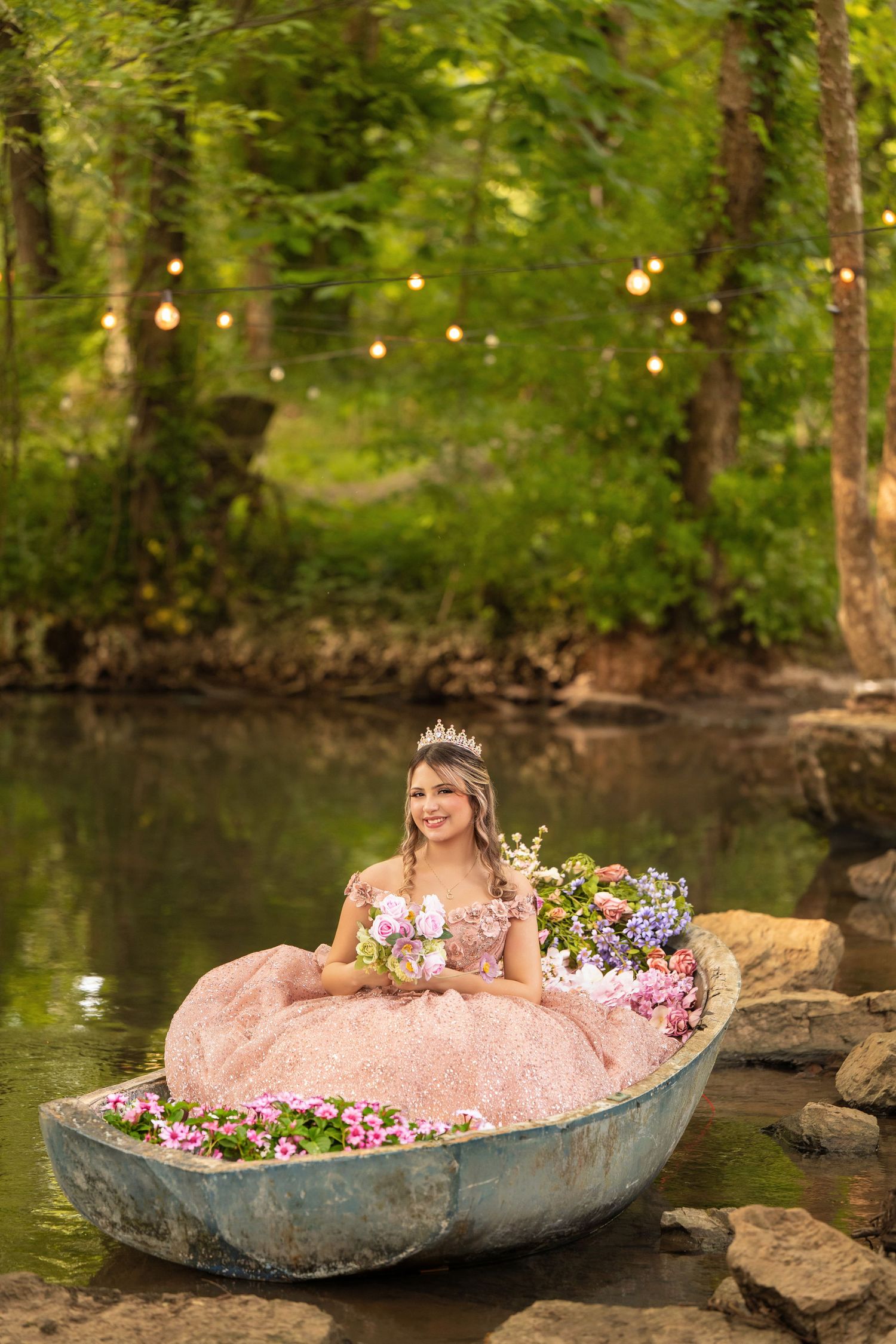 Girl in pink ball gown with tiara sits in flower-filled boat on serene woodland pond with string lights.