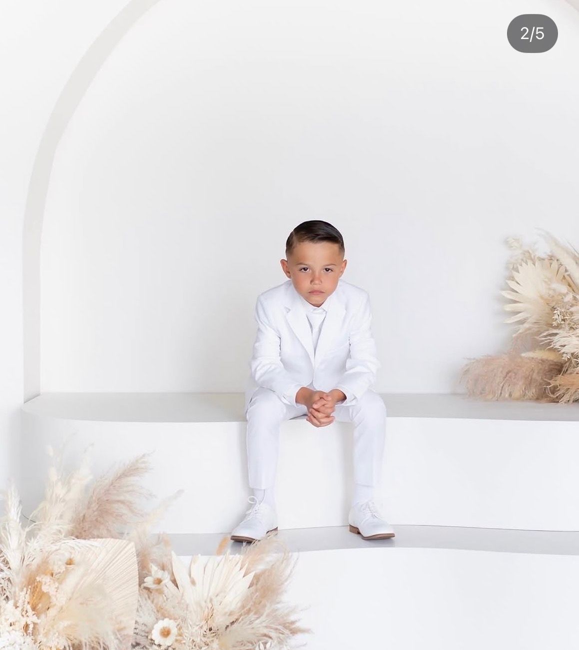 Young boy in a white suit sits on a white ledge surrounded by dried pampas grass in a bright, minimalist setting.