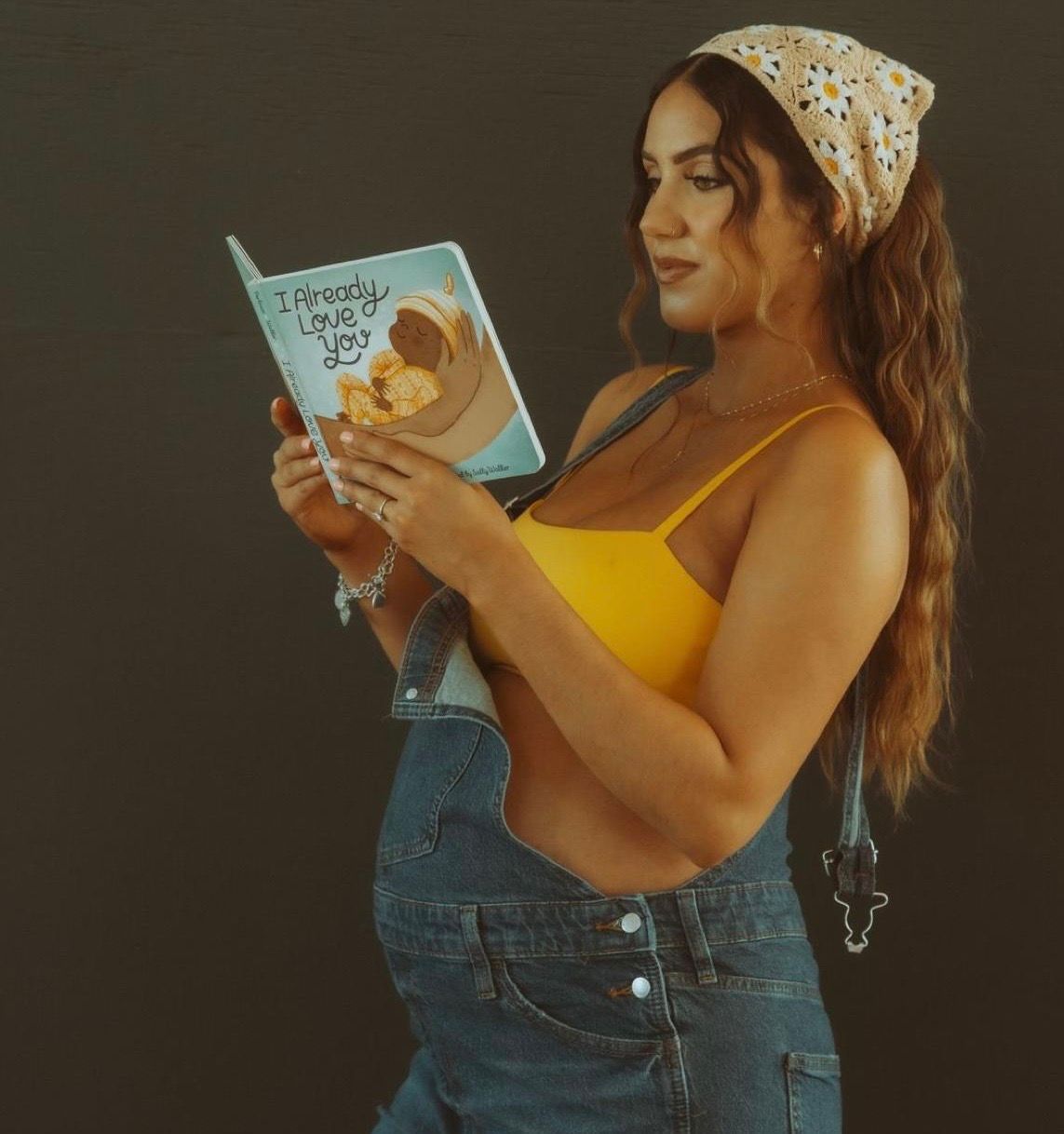 Pregnant woman in yellow crop top and denim overalls reading a baby book titled I Already Love You.