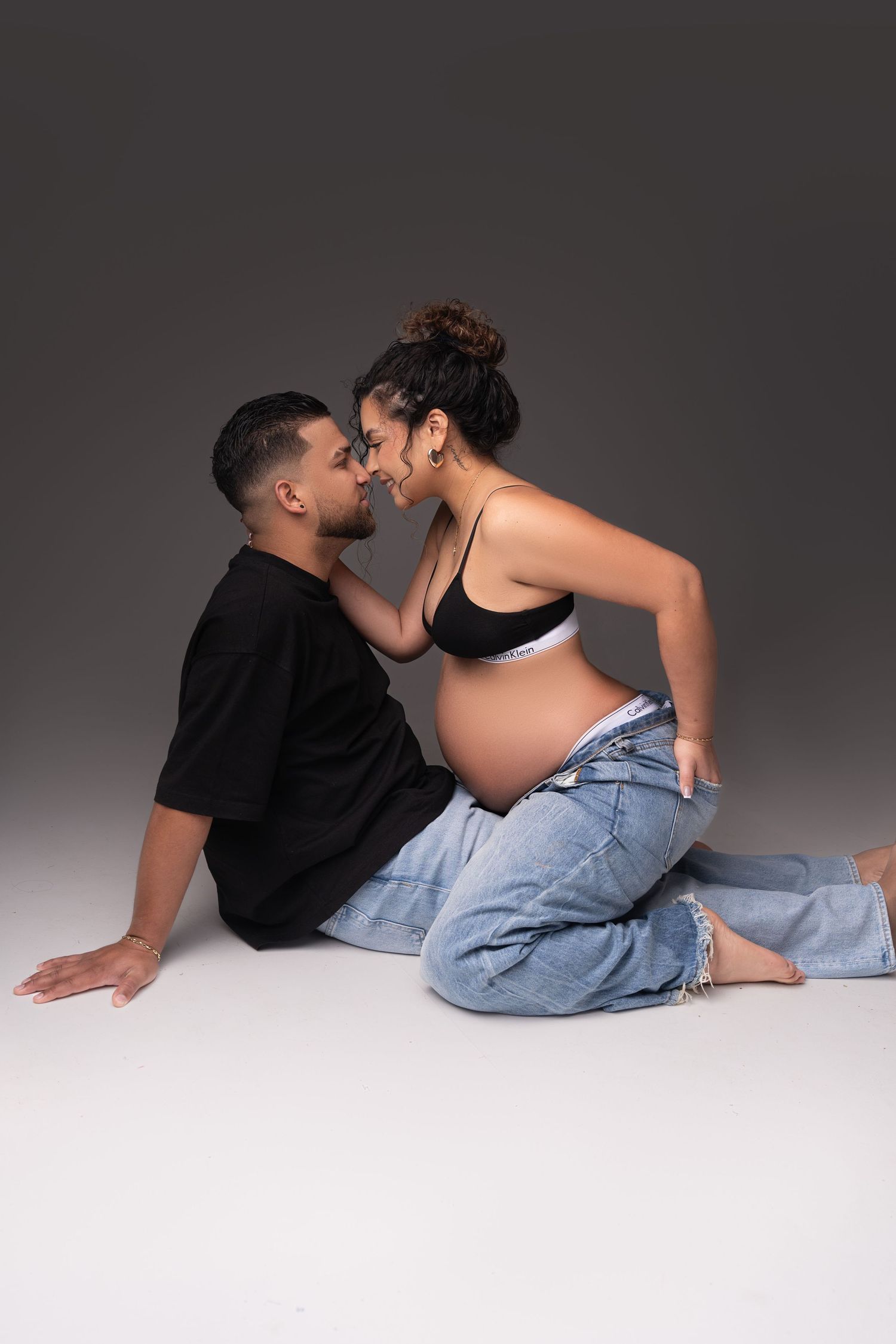 A couple in casual attire lying close together on a gray studio floor in a romantic photoshoot sequence.