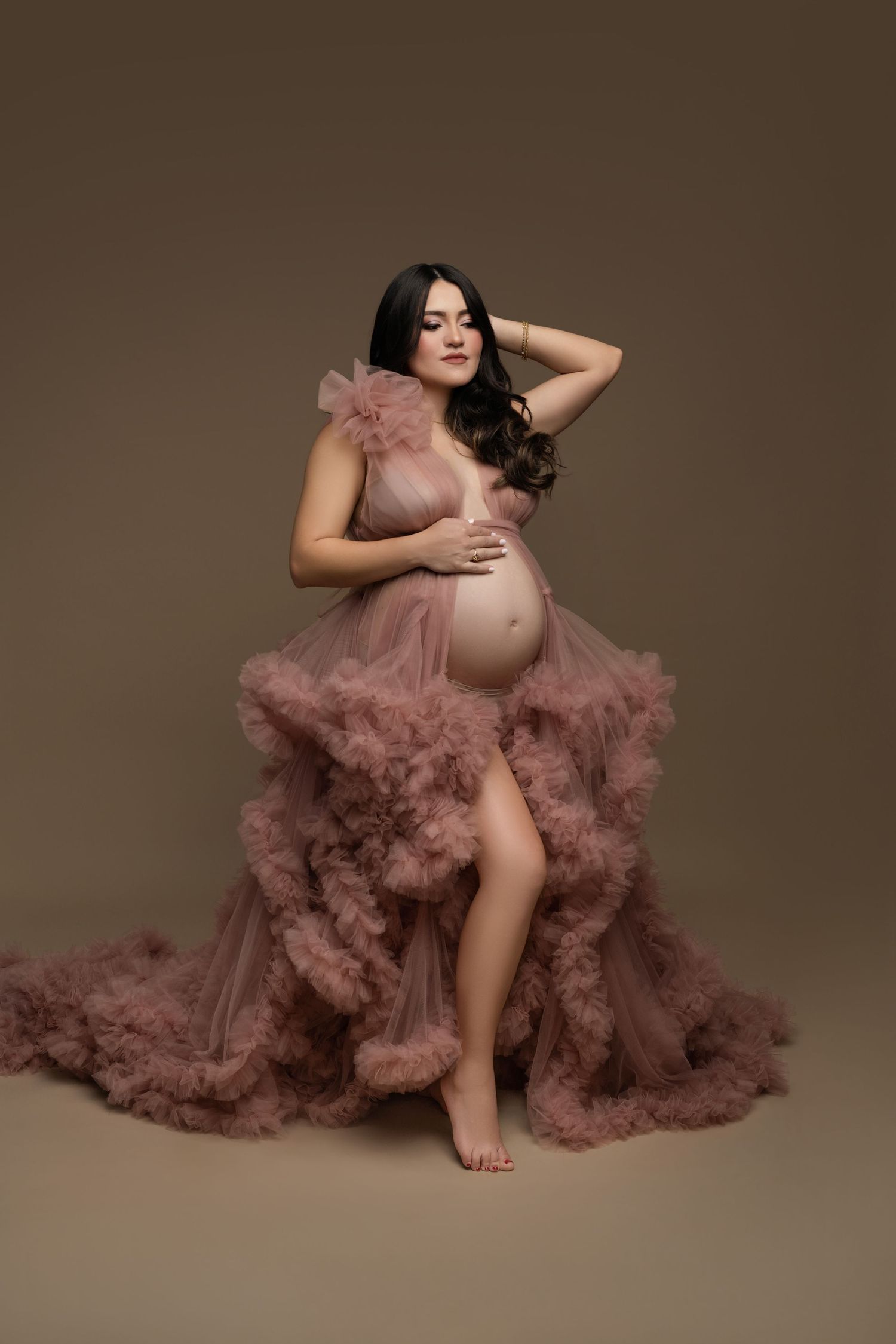 A figure in a flowing dusty rose tulle gown poses gracefully against a neutral studio backdrop in a maternity photoshoot.