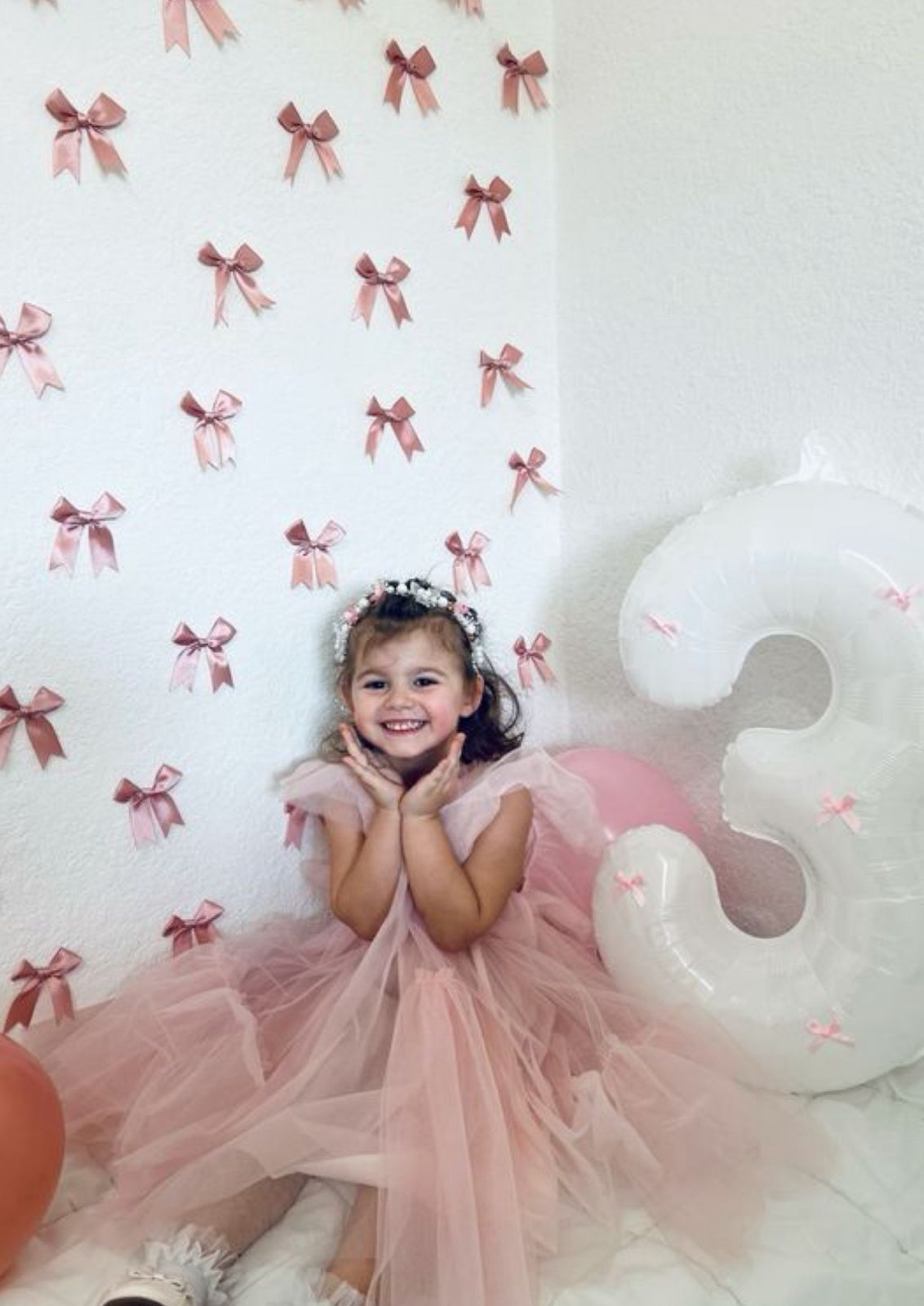 A smiling girl in a pink tutu dress poses by a bow-decorated wall next to a number 3 balloon at her birthday party.