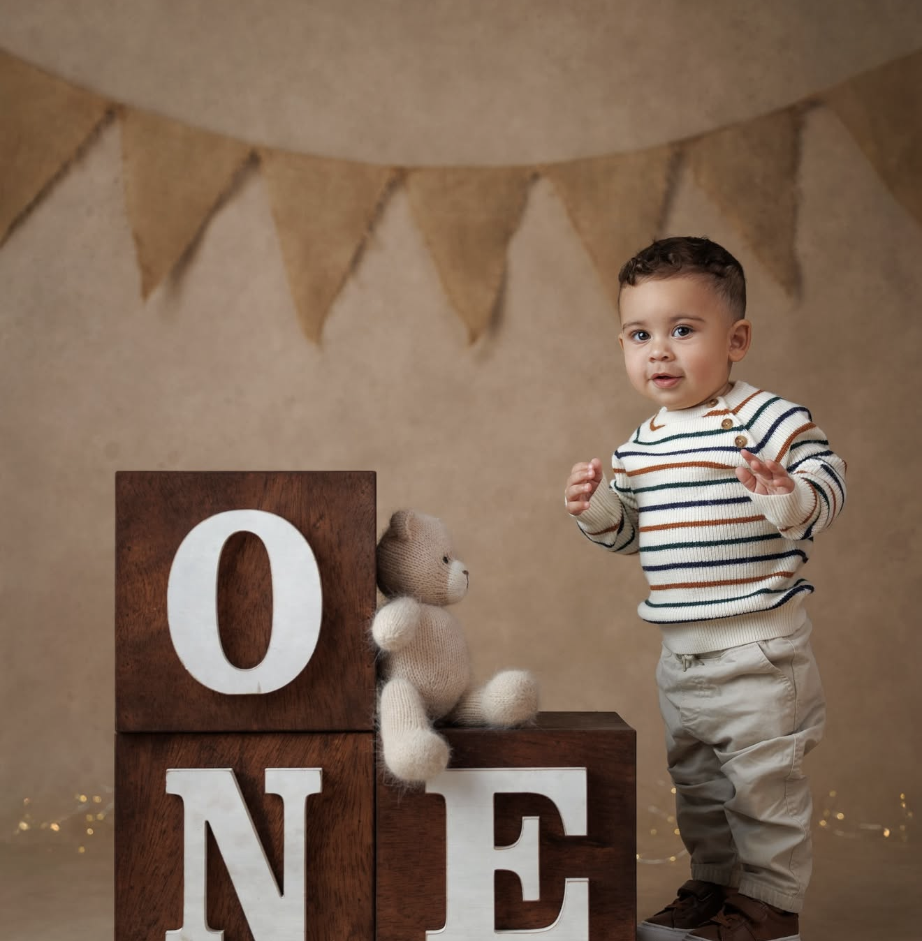 Toddler boy in striped shirt poses beside wooden ONE blocks with teddy bear for first birthday photo.