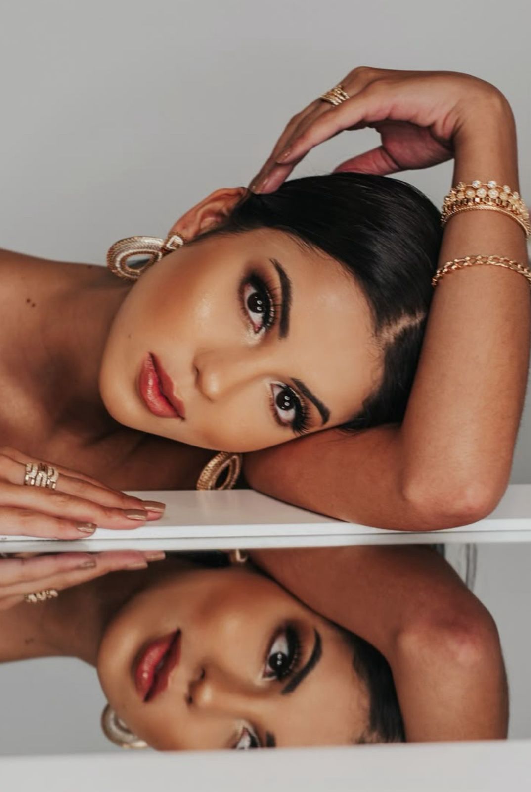 Woman with dark hair poses at mirror wearing gold jewelry, rings, and bracelets, with reflection visible below.