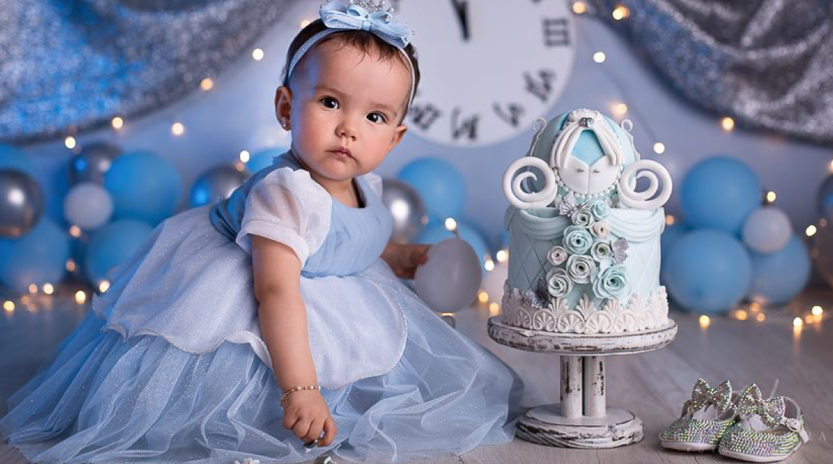 Baby girl in blue tulle dress sits beside a Cinderella-themed first birthday cake with balloon decorations.