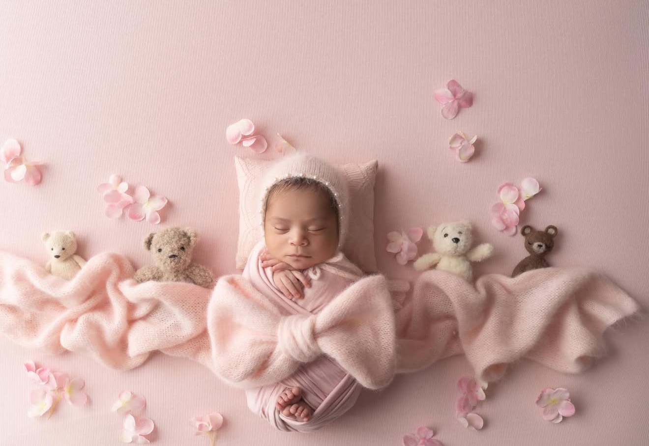 Newborn baby in pink bonnet wrapped in pink blanket surrounded by tiny teddy bears and flowers on pink background.