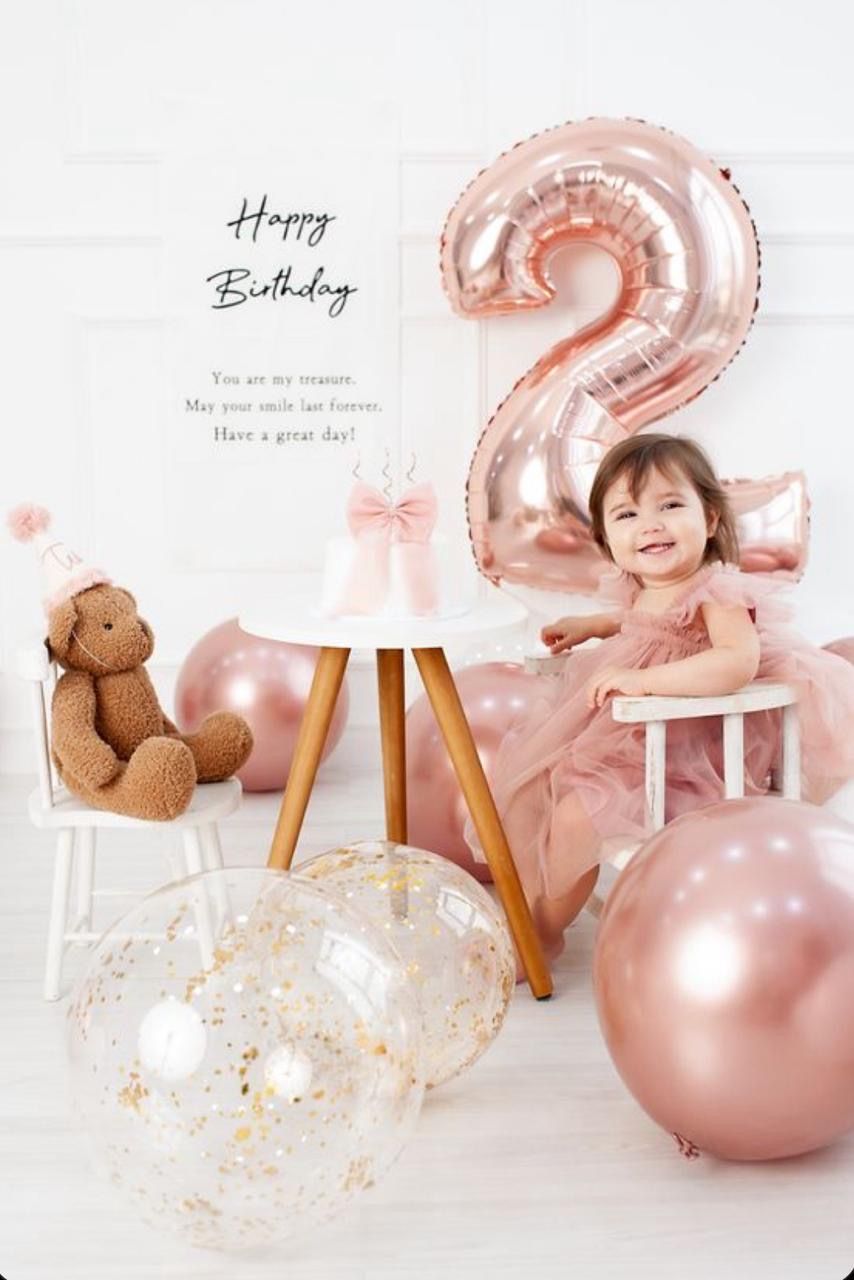 Toddler girl in pink dress celebrating 2nd birthday with rose gold balloons, teddy bear, and Happy Birthday sign.