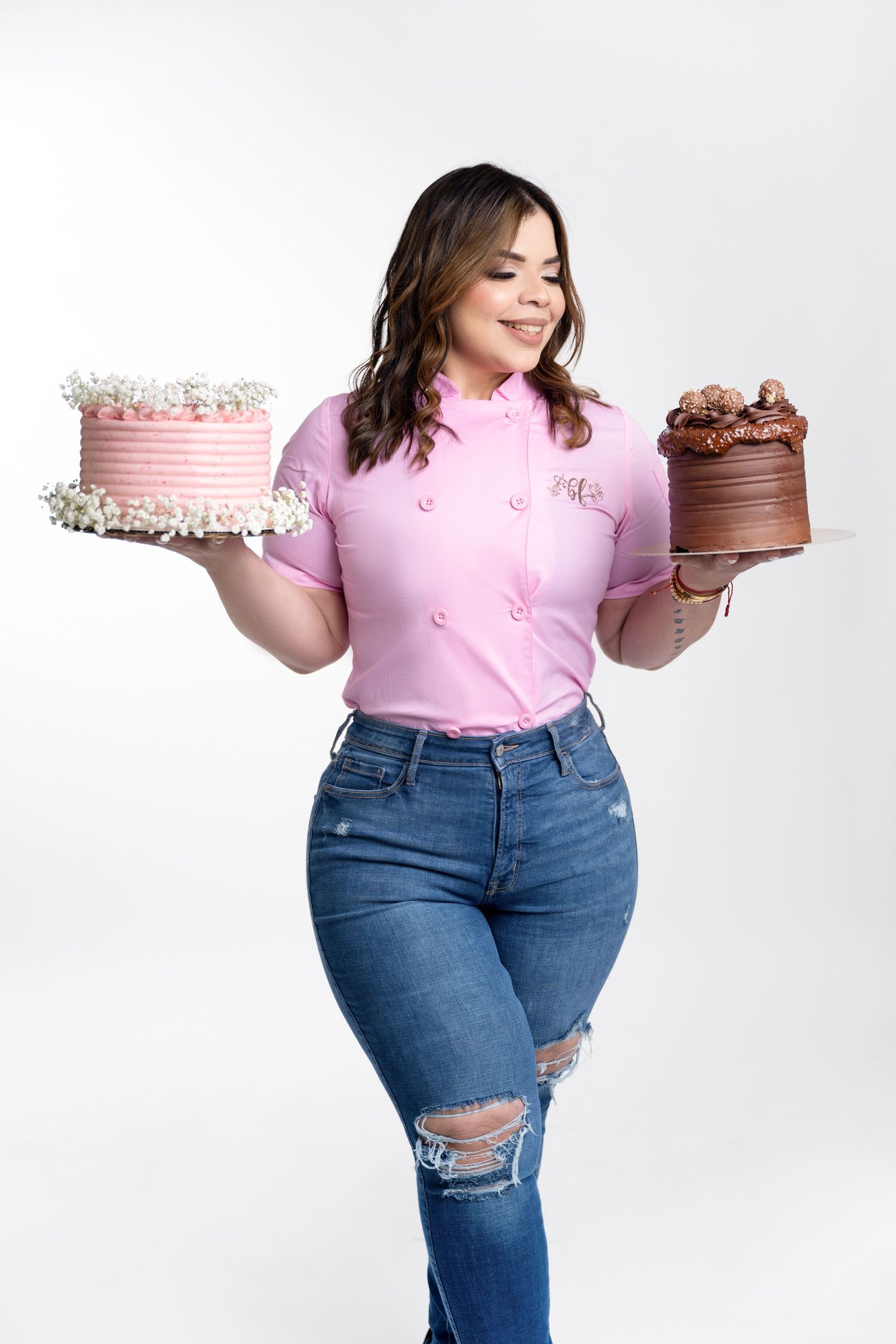 Smiling woman in a pink chef shirt and ripped jeans holds a pink frosted floral cake and a chocolate cake against a white background.