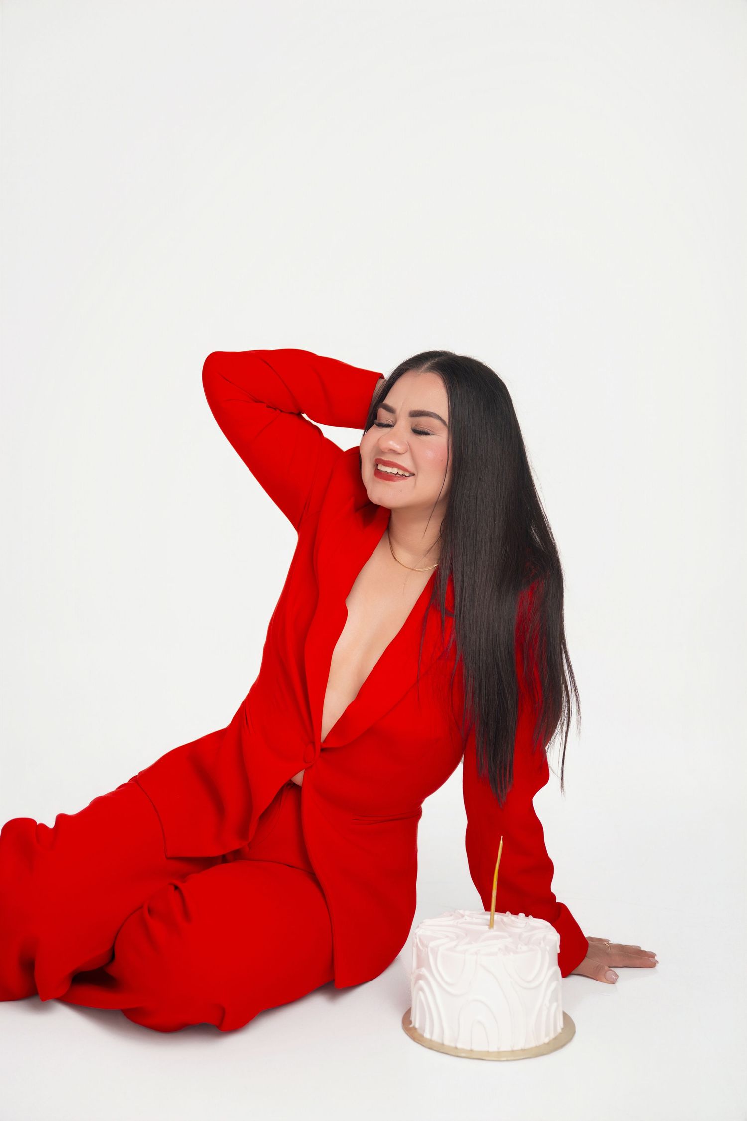 Smiling woman with long dark hair in a red blazer suit sits on a white floor with a hand behind her head beside a small white birthday cake.