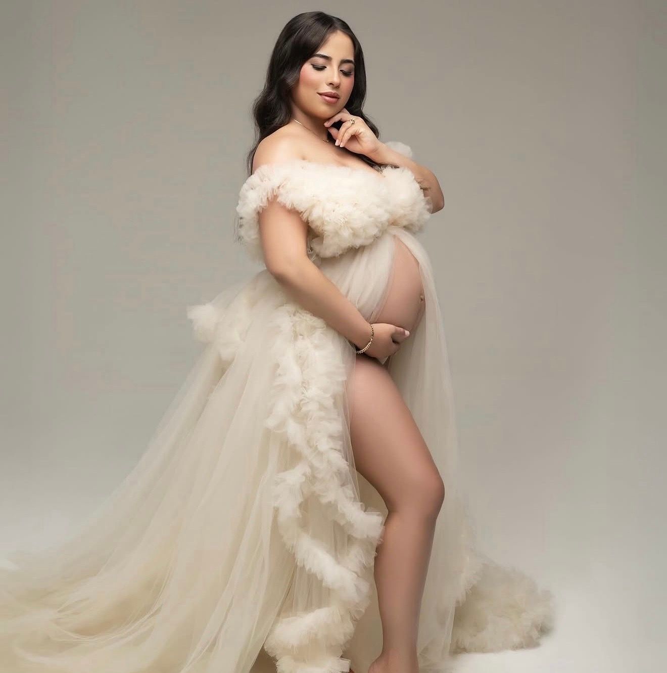 Pregnant woman in a cream off-the-shoulder tulle maternity gown with ruffles posing in a studio and cradling her baby bump.