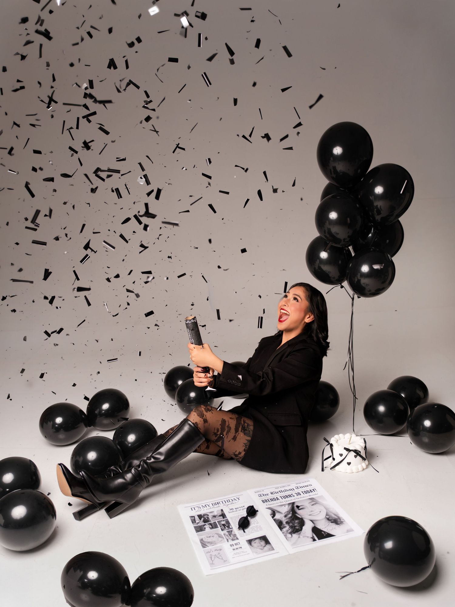 Woman laughing sitting on floor surrounded by black balloons and confetti, holding champagne bottle in monochrome celebration setup.