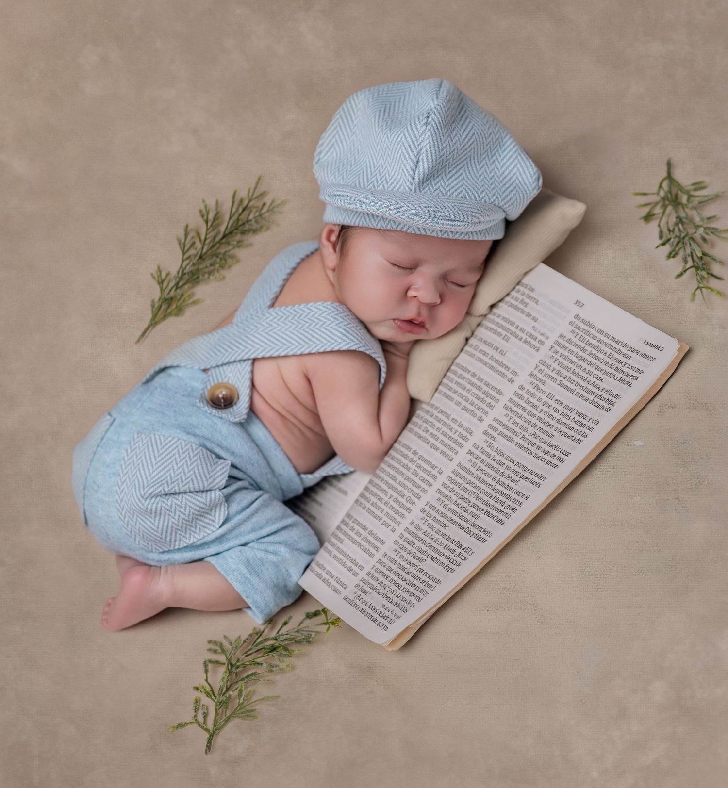 Sleeping newborn baby wearing a light blue knit cap and overalls, resting on an open book with rosemary sprigs.