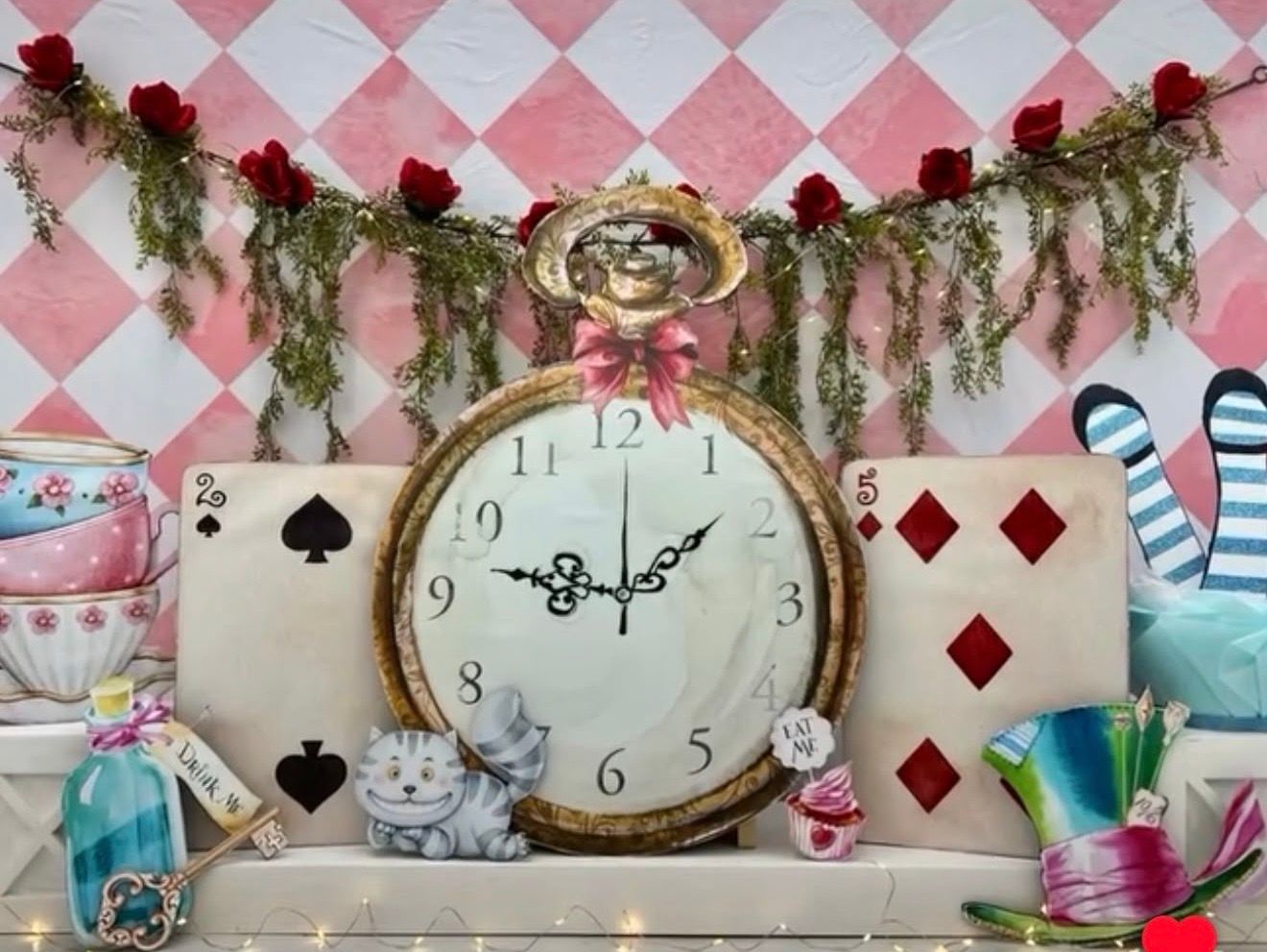 Alice in Wonderland themed display with large clock, playing cards, Cheshire cat, and red roses on pink checkered background.