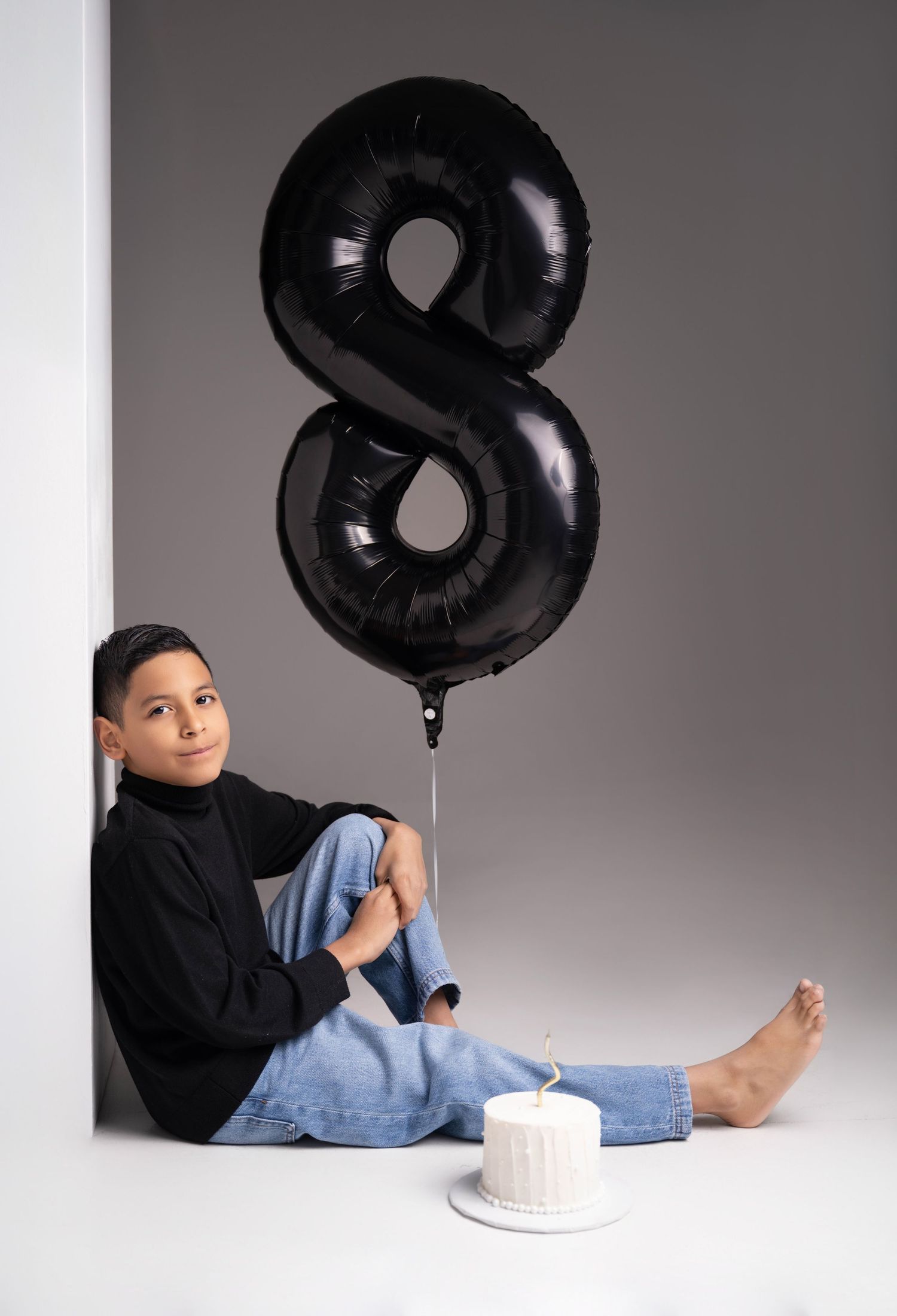 A boy sits against a wall with a black number 8 balloon and a white birthday cake, celebrating his 8th birthday.