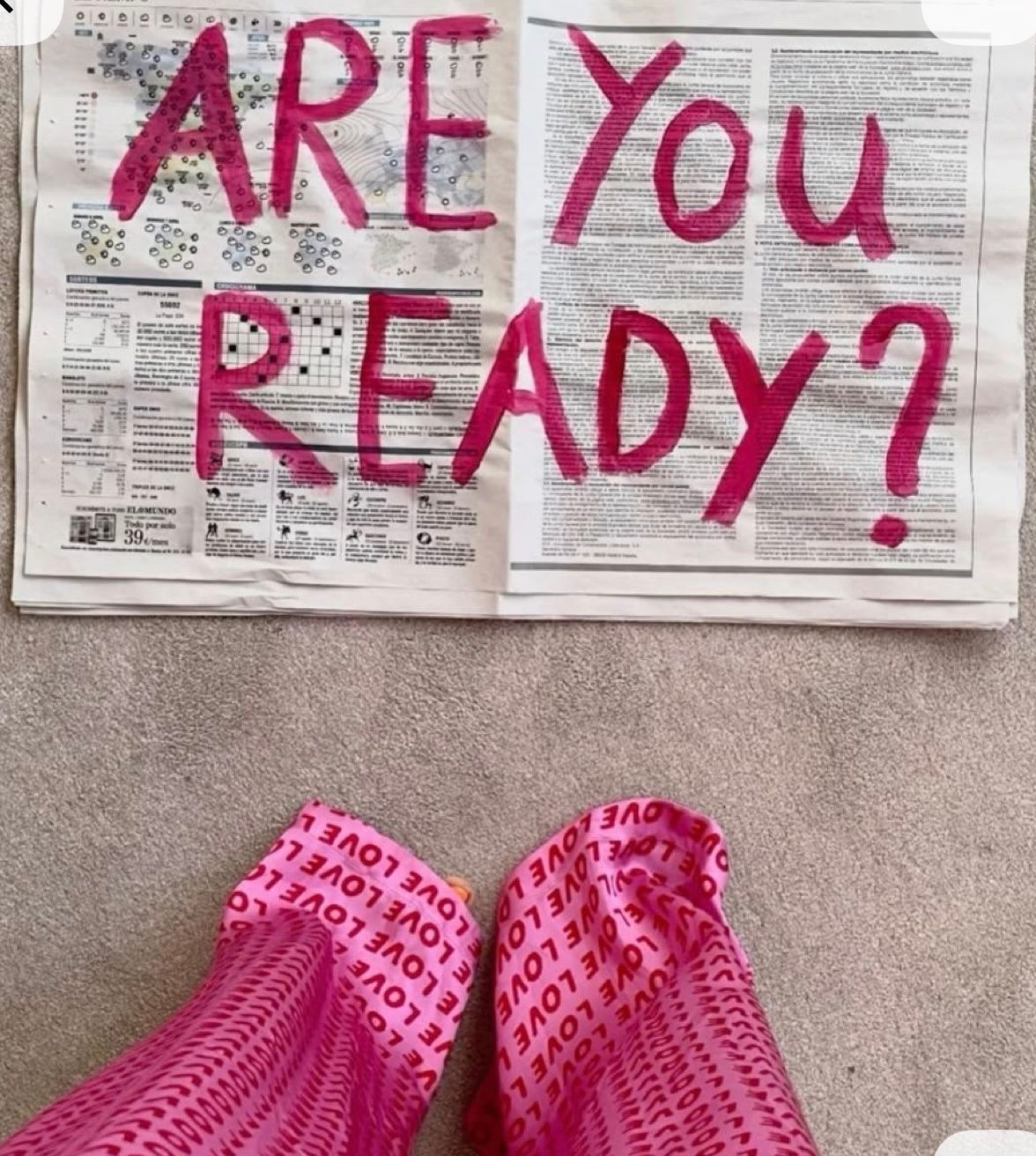 Pink socks with LOVE text next to newspaper with ARE YOU READY written in pink paint on carpet floor.