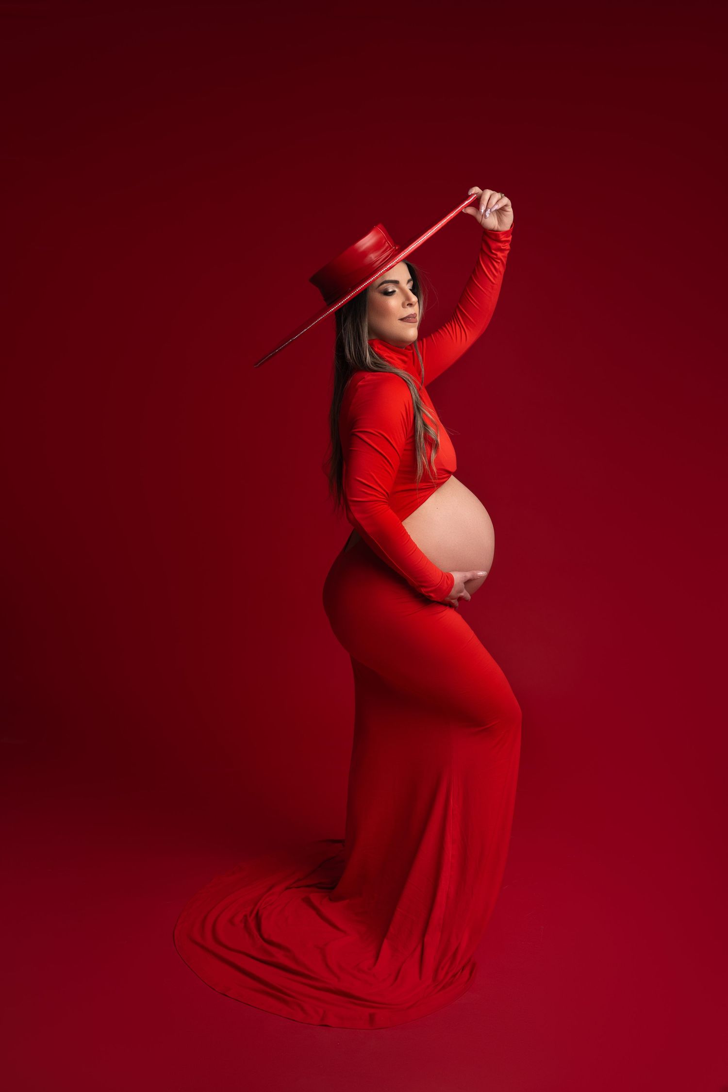 Woman in all red attire with hat poses for maternity photo against red background.