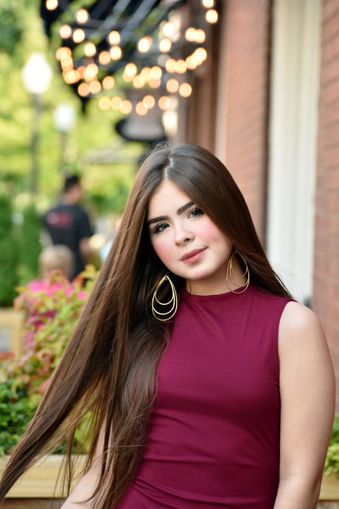 Portrait in burgundy sleeveless dress with long straight hair and statement earrings.