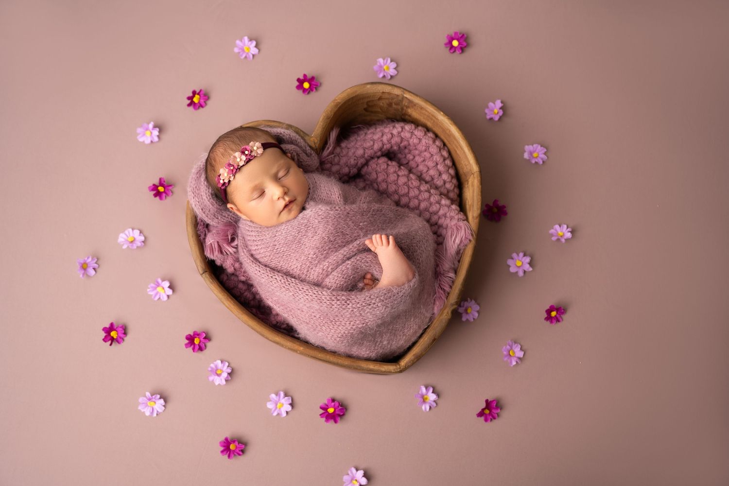 Sleeping newborn in purple wrap surrounded by pink flowers in heart-shaped basket.