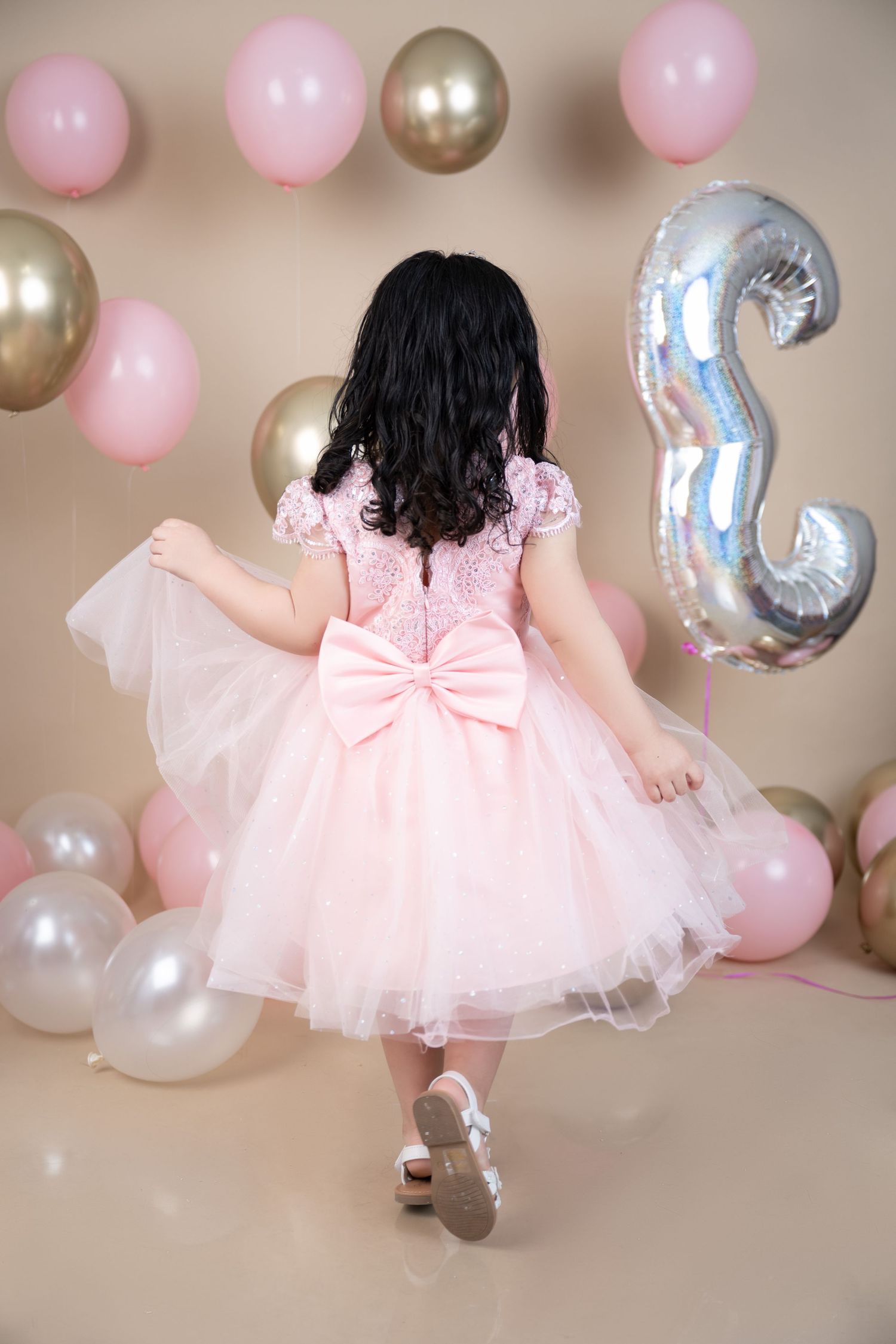 Pink and gold balloons decorate backdrop as someone twirls in fluffy pink tulle dress with large bow.