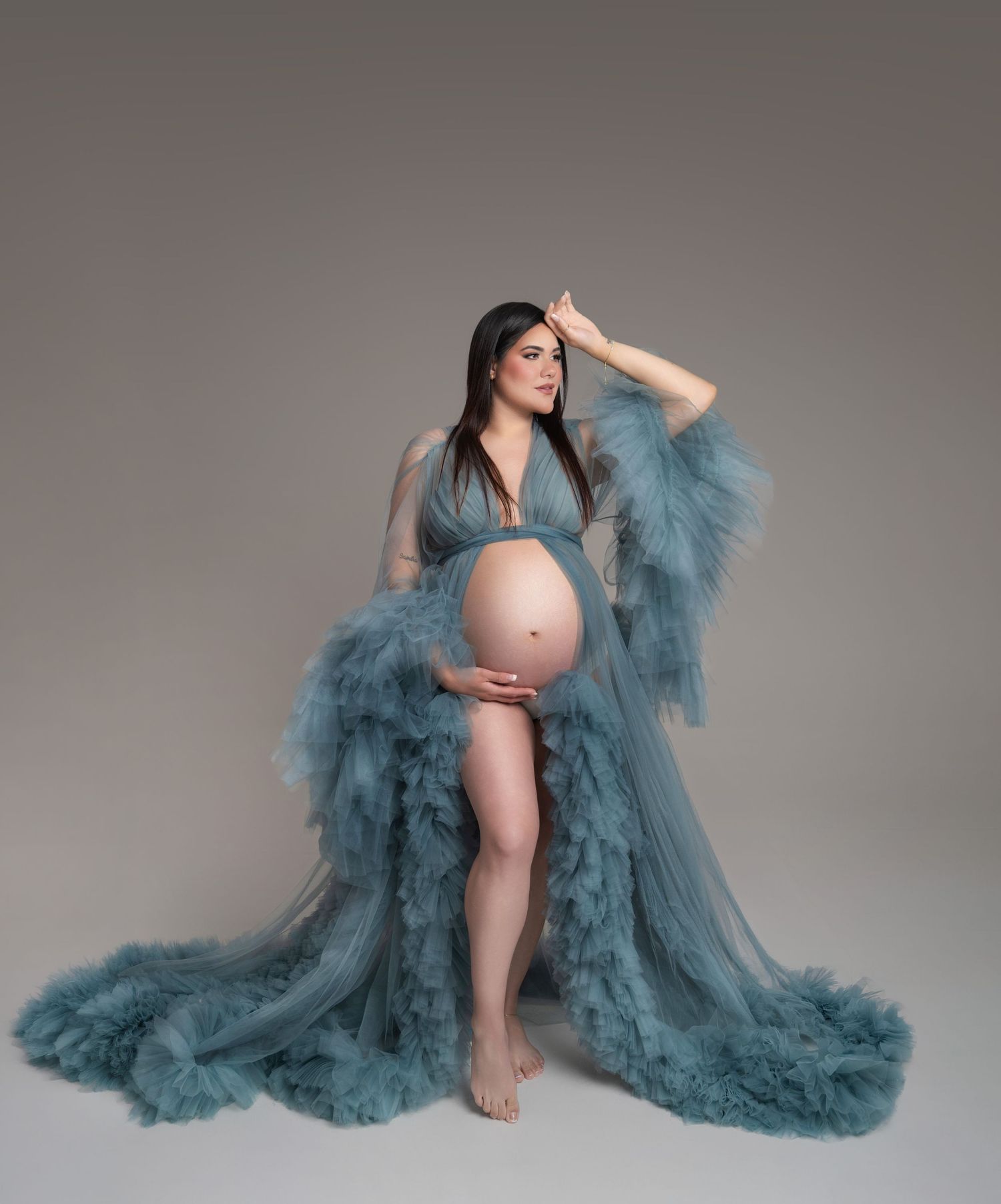Pregnant woman in flowing teal tulle maternity robe posing gracefully in studio, showcasing baby bump.