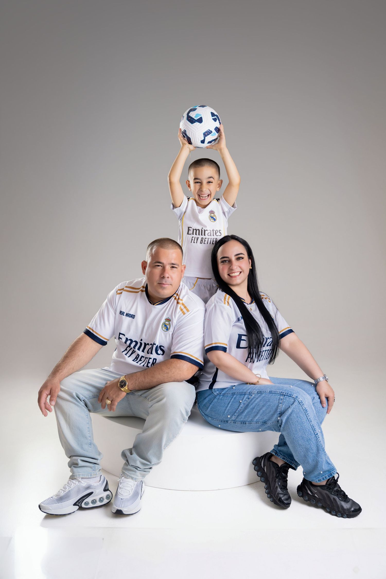 Family of three in matching Real Madrid jerseys, parents sitting with child standing behind holding a soccer ball.