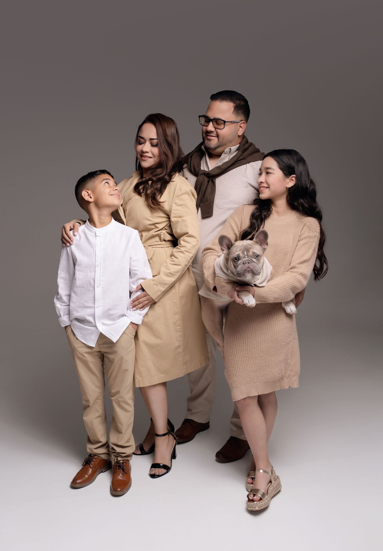 Family of four in coordinated beige outfits posing together in studio, with girl holding French Bulldog puppy.
