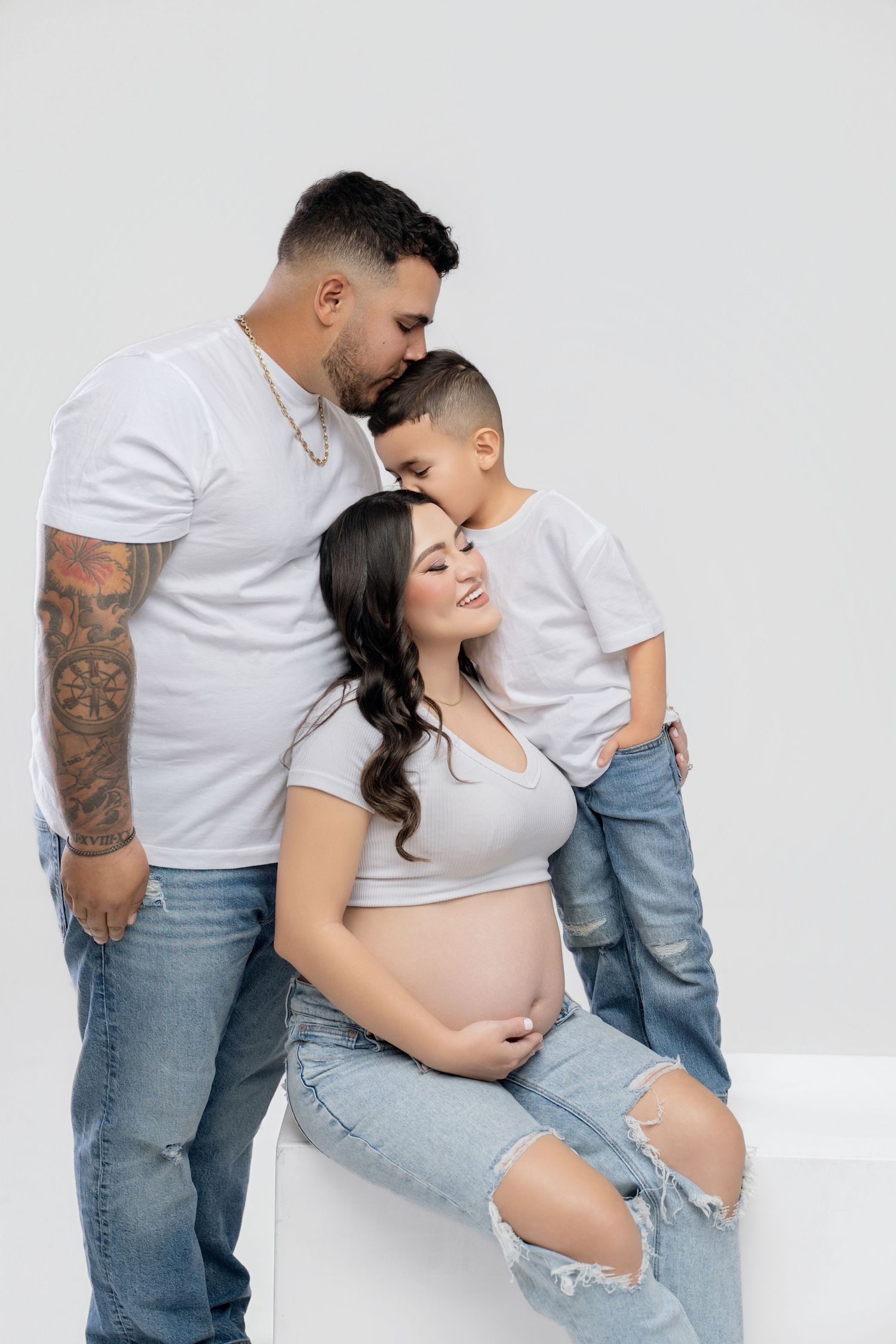 A tattooed man and young boy kiss a smiling pregnant woman in white tops and jeans against a white background.