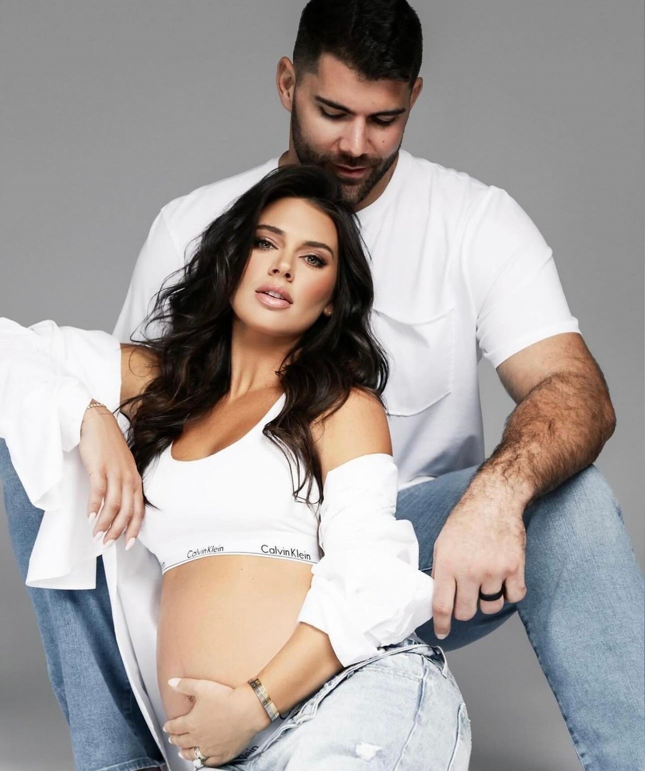 Couple posing in Calvin Klein underwear and white outfits, woman showing baby bump in maternity photo shoot.