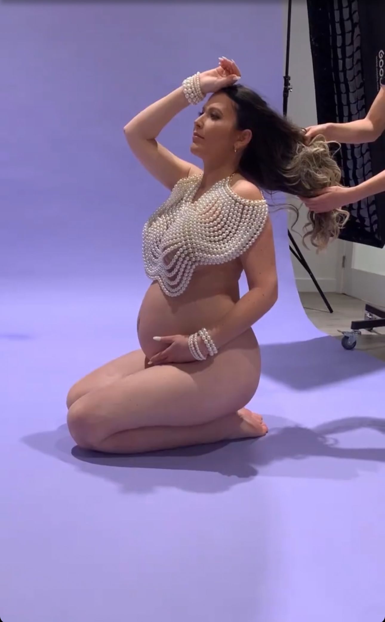 Pregnant woman in pearl bodysuit kneels gracefully during a professional maternity photo shoot.