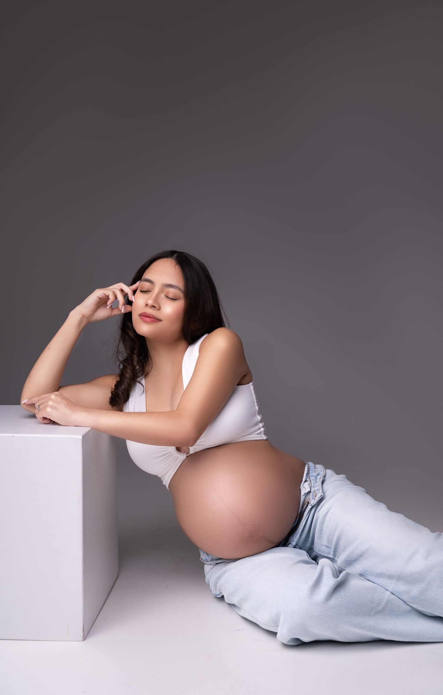 A pregnant woman in white top and light jeans poses elegantly against a gray studio backdrop in three artistic maternity photos.