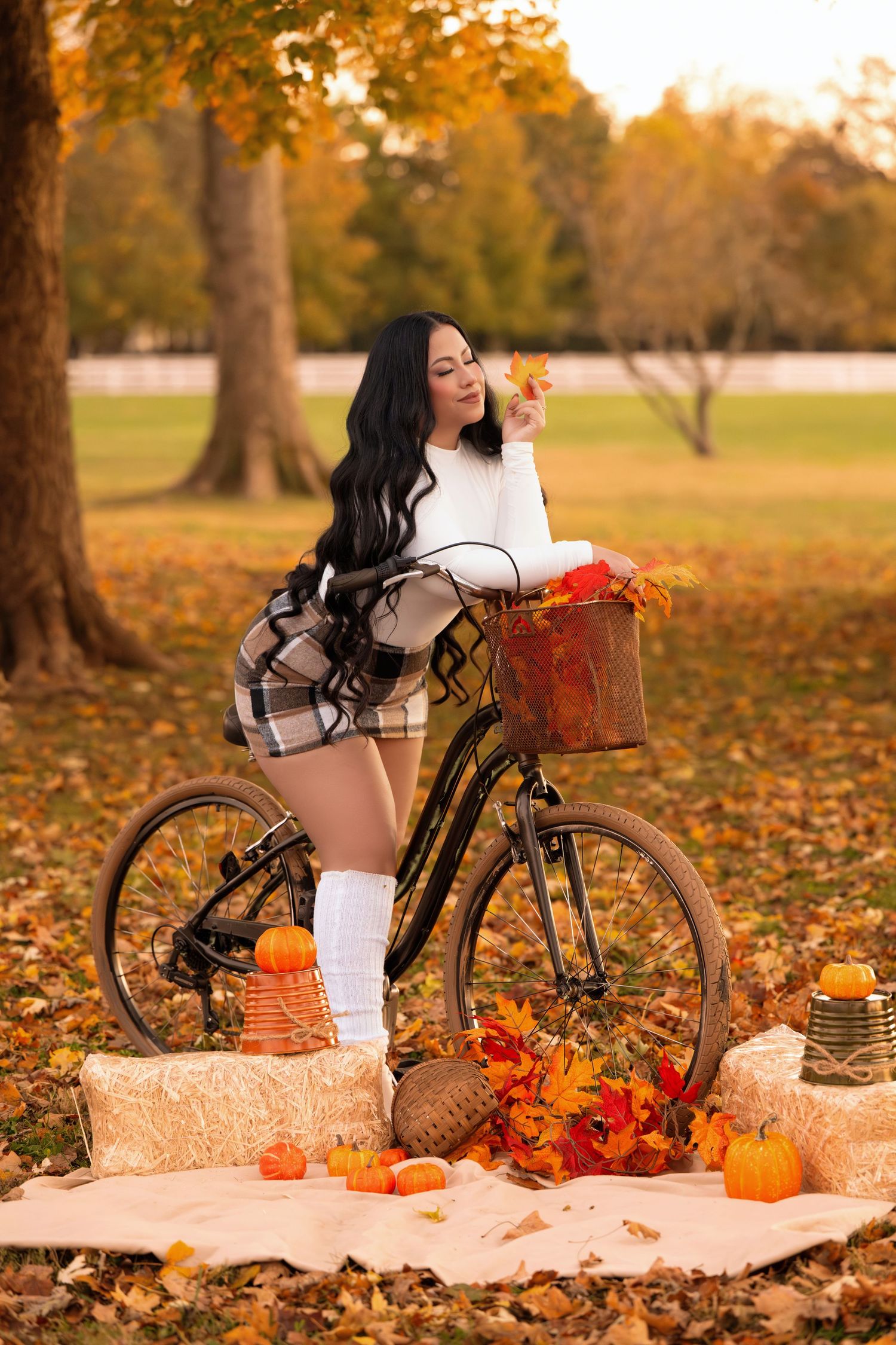 Woman in plaid skirt and white sweater posing with vintage bicycle among fall pumpkins and autumn leaves in park.