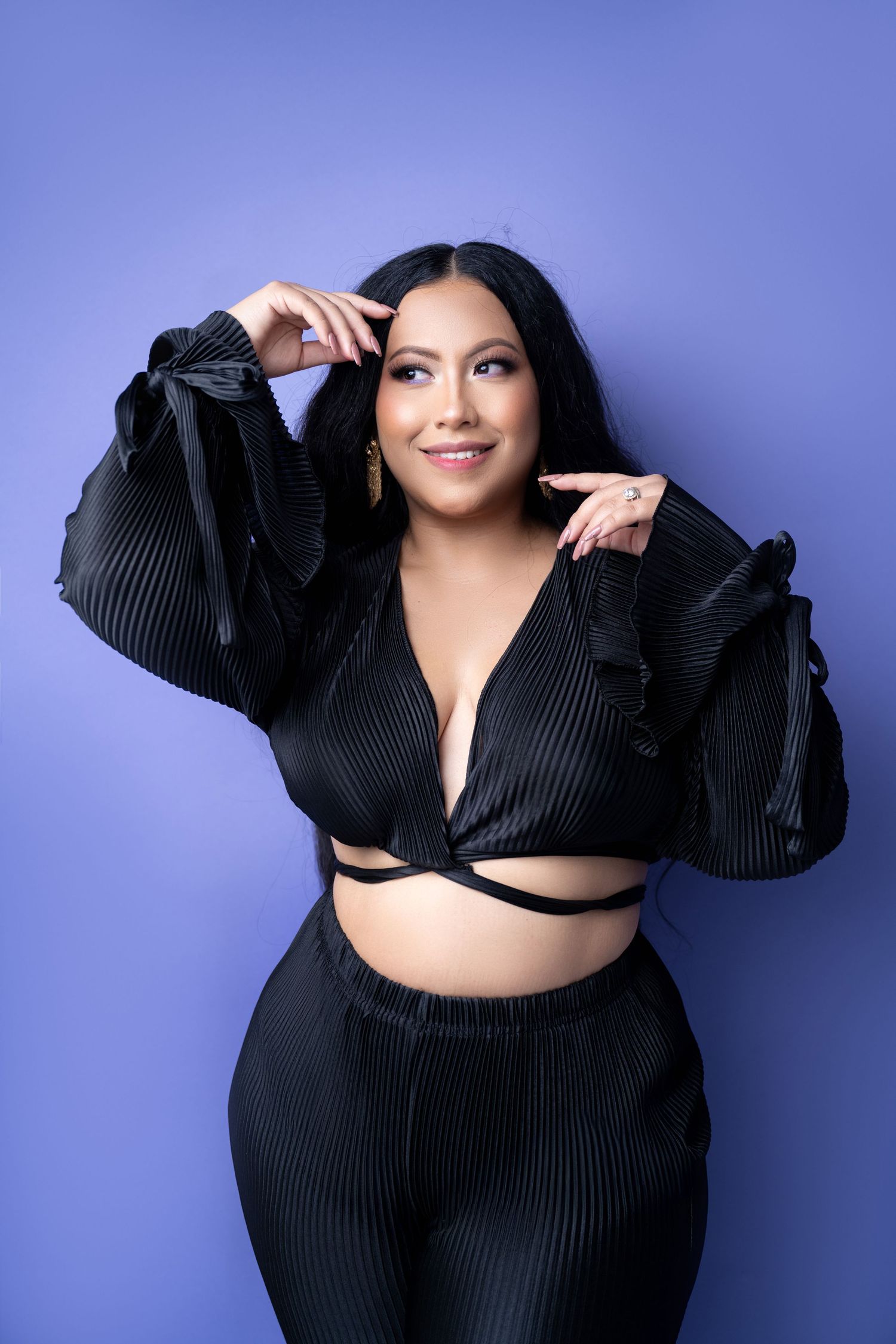 Woman with long black hair poses against a purple backdrop in a black pleated crop top and matching pants, hands raised near her face.