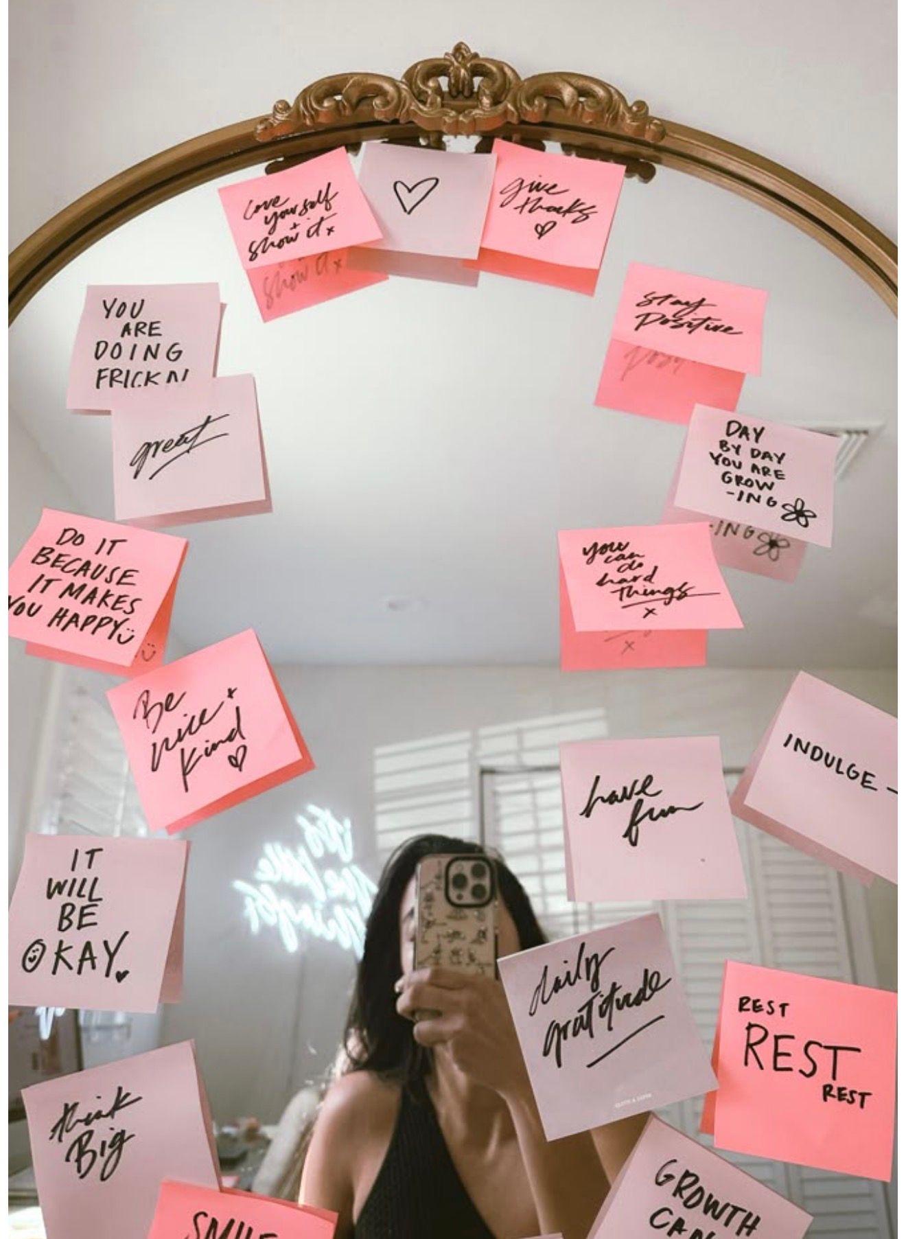 Woman taking mirror selfie surrounded by pink sticky notes with positive affirmations and motivational quotes on a gold mirror.