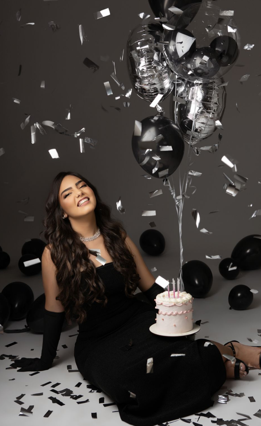 Woman in black dress sits on floor with birthday cake, surrounded by black balloons and silver confetti falling.