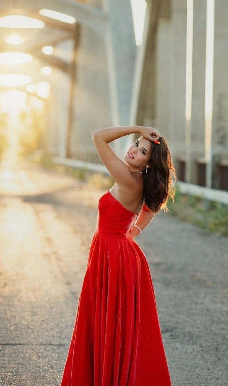 Woman in elegant red gown poses gracefully under bridge in golden sunset light.