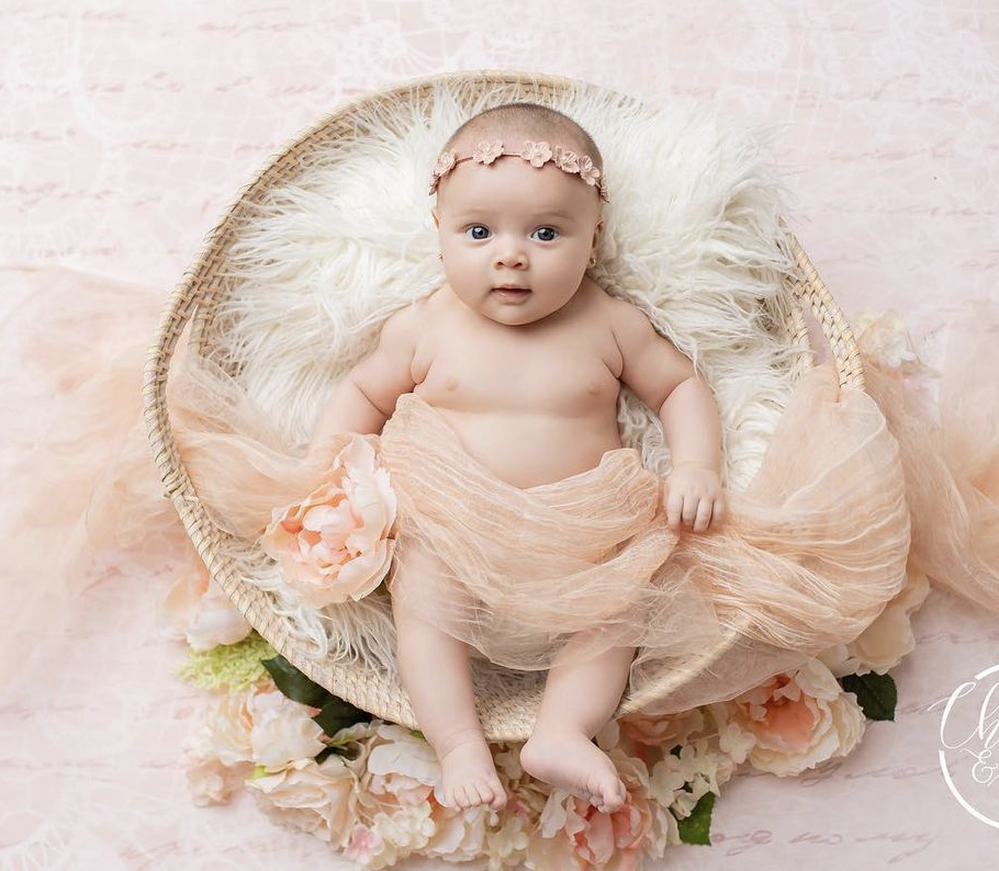 Newborn baby girl in peach tutu sitting in wicker basket with white fur and flowers.