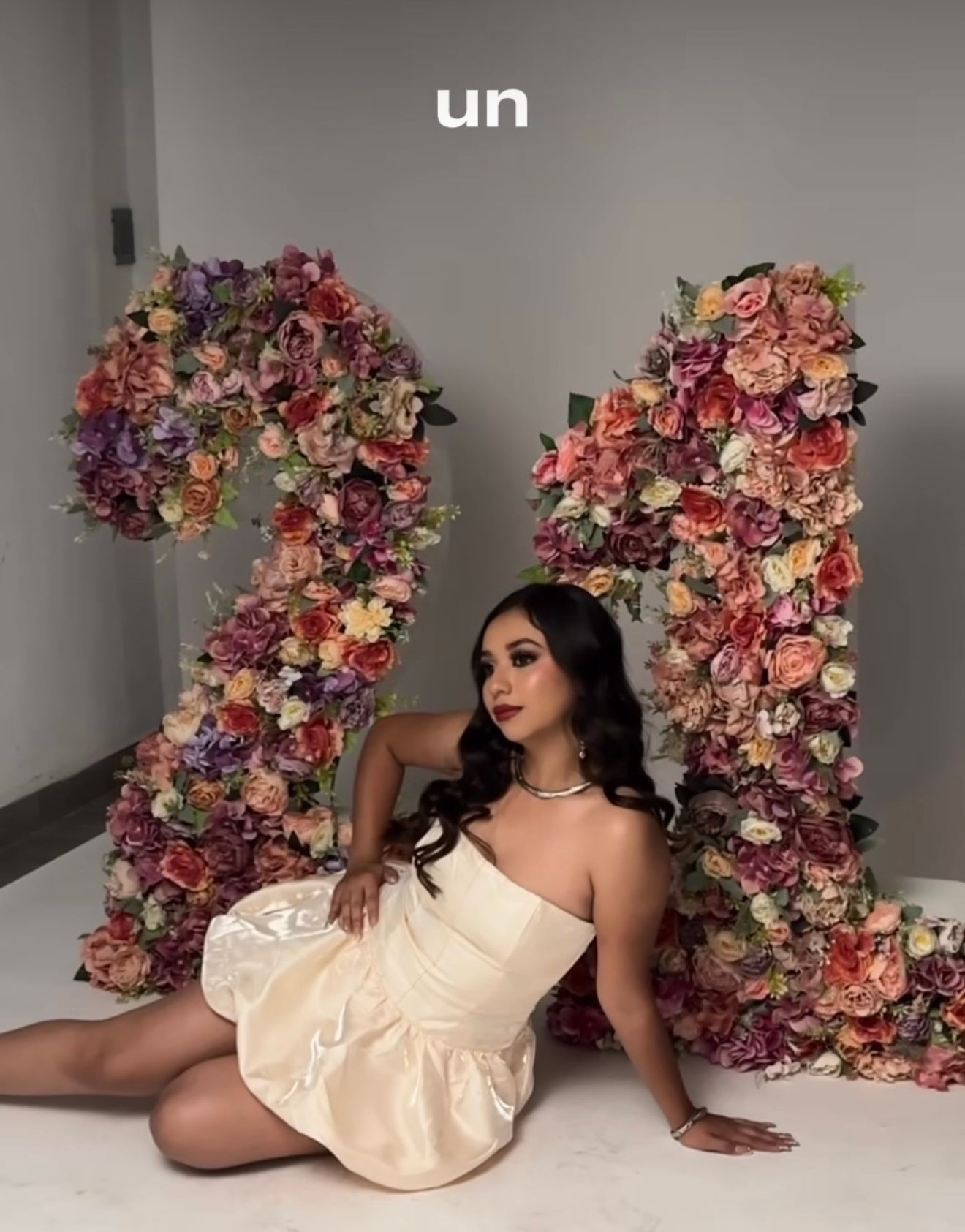 Young woman in white dress poses between large floral number 21 decorations for a birthday celebration.