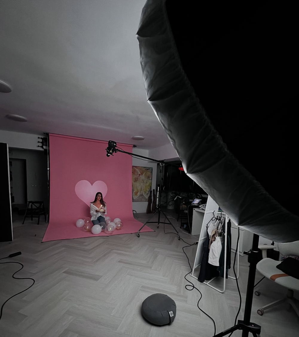 A photographer's studio with a pink backdrop, heart balloon, and a woman posing during a professional photo shoot.
