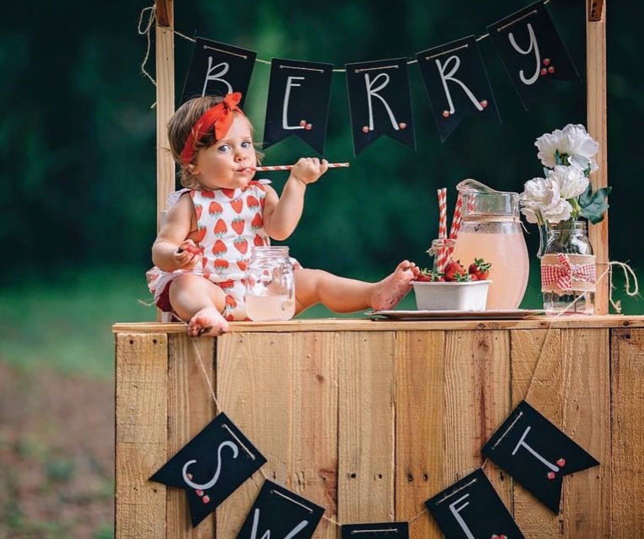 Adorable baby girl sitting on rustic wooden berry sweet stand, wearing strawberry romper with red bow headband outdoors.
